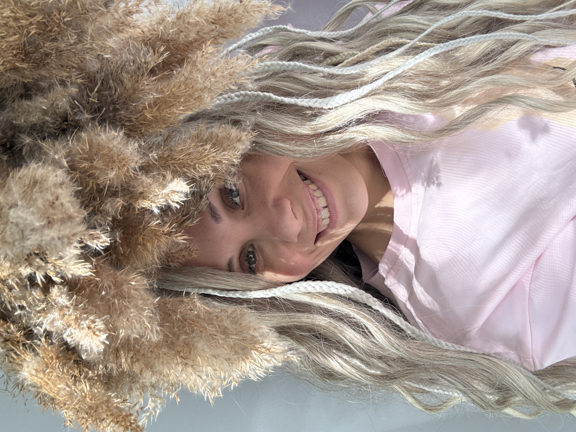 The image depicts a woman with blonde hair and white braids, lying down against a light-colored surface. Her face is angled towards the viewer and she is smiling. She is wearing a light pink shirt. To her left and above her head is a collection of dry, feathery plants, possibly pampas grass. The setting appears to be indoors, given the presence of an artificial light source creating shadows. The location of Rusanovo, Russia is not discernible from the image.