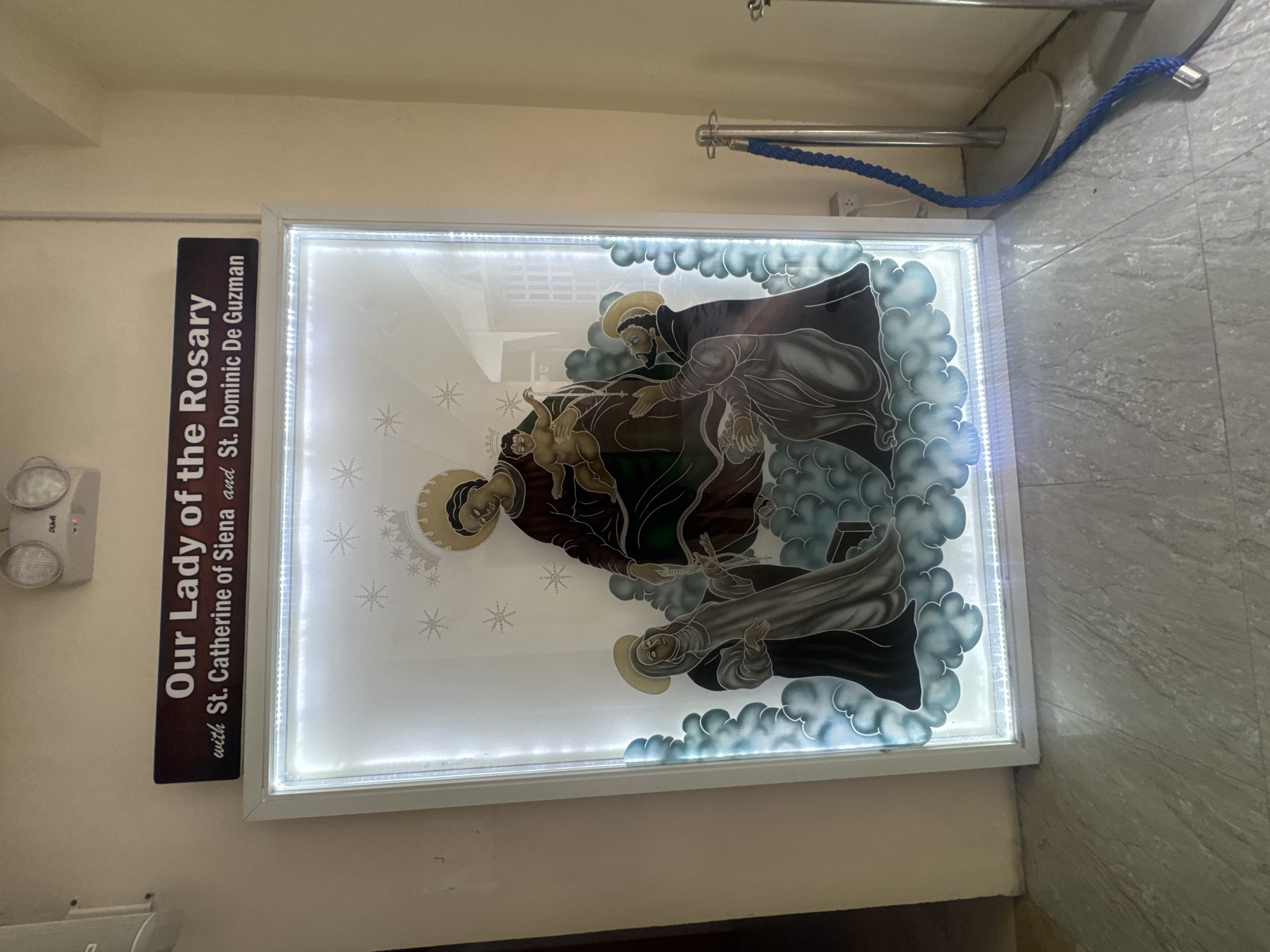 The image shows an illuminated glass artwork titled "Our Lady of the Rosary with St. Catherine of Siena and St. Dominic De Guzman". The artwork depicts the Virgin Mary holding the infant Jesus, with St. Catherine of Siena and St. Dominic De Guzman on either side, all appearing to be floating on clouds. The artwork is framed in white and lit from within, creating a halo effect around the figures. Above the artwork is a dark rectangular sign with white text. The artwork is displayed indoors, on a light-colored tiled floor, with a blue rope barrier and stanchions to the right. The background wall is a light beige color, and a white emergency light fixture is visible on the ceiling. This artwork is located at the Ateneo de Davao University, a Jesuit university in Davao City, Philippines.