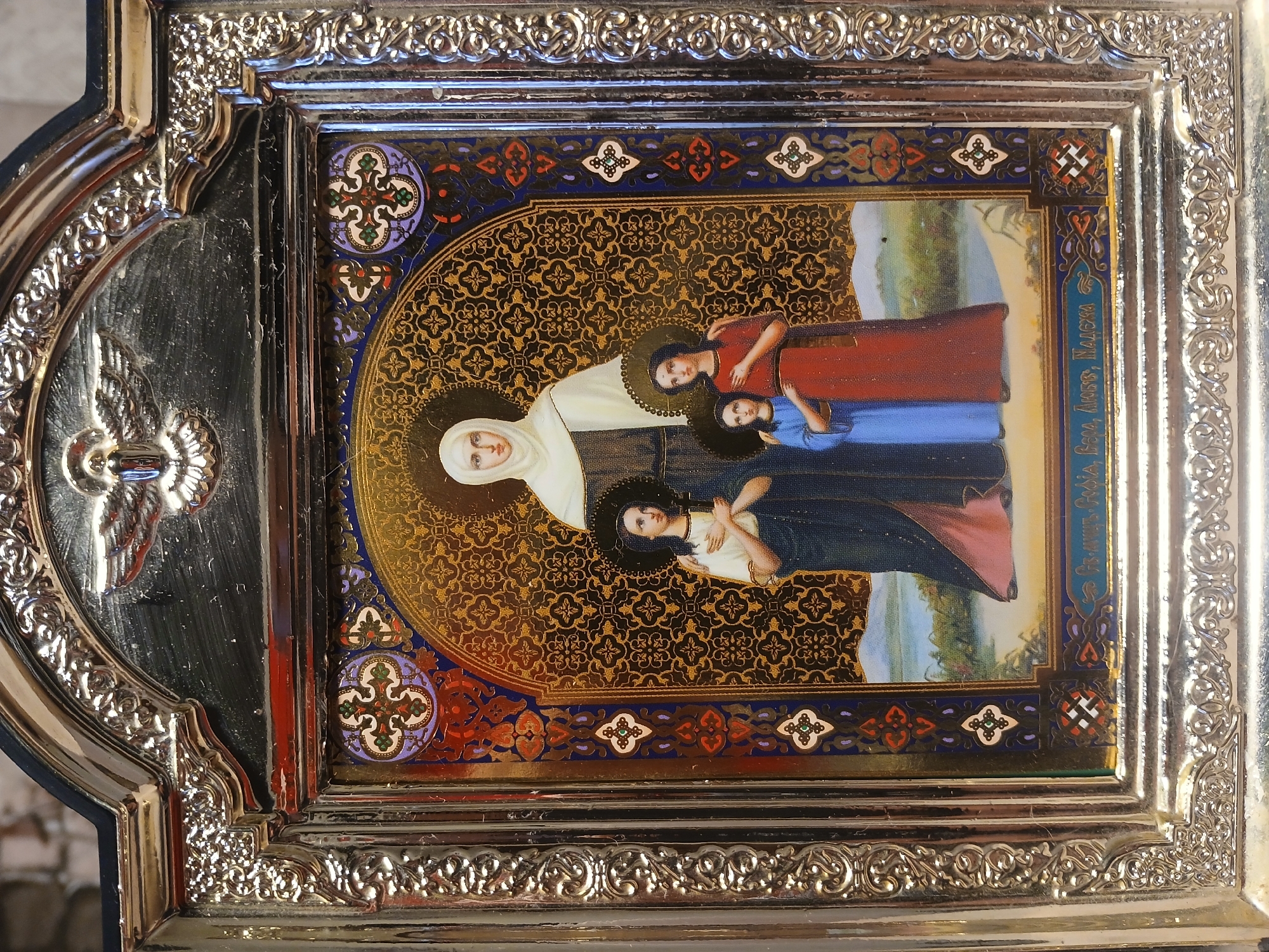 The image shows a framed icon, likely of Eastern Orthodox origin. It depicts three female figures with halos, the central one in white robes, and the other two in colored robes. The icon has intricate decorative borders with geometric and floral patterns. The frame is silver with ornate embellishments. The location is Kamianske, Ukraine.