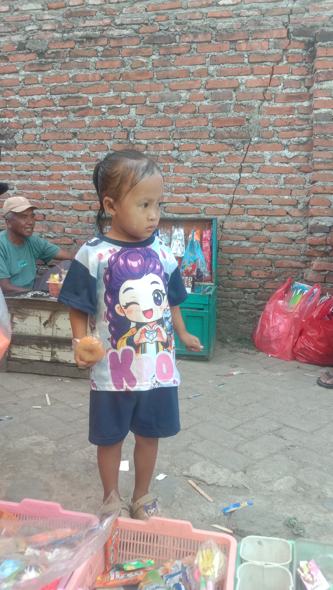 A young girl stands in front of a brick wall in Karang Anyar, Indonesia. She is wearing a t-shirt with a cartoon character and dark blue shorts. In her right hand, she holds a small, round snack. To her left, a man in a tan cap and green shirt sits behind a wooden cart. Behind the girl, a green cabinet displays various items, and red plastic bags are visible to the right. The ground is paved with concrete blocks and scattered with small wooden sticks. Two pink baskets filled with packaged snacks and other items are in the foreground.