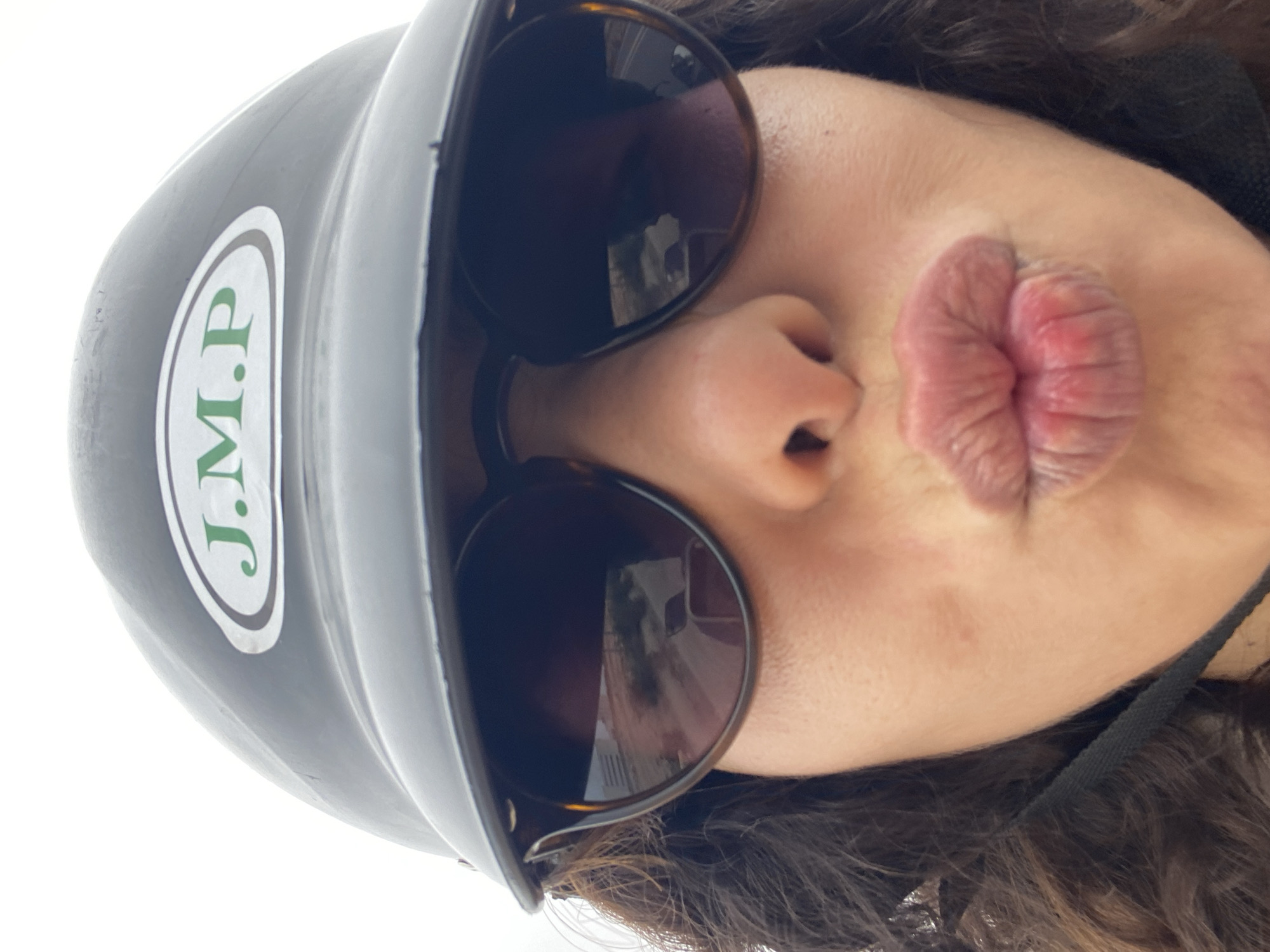 A selfie is taken from a low angle, showing a person's face from just above the eyes to the mouth, with a helmet on. The helmet is dark grey with a white oval logo containing green text that reads "J.M.P.". The person is wearing dark sunglasses, and their lips are pursed in a kissing gesture. The background is bright white and out of focus. The reflection in the sunglasses shows what appears to be a street scene with buildings and possibly a vehicle. The subject's curly, dark hair is visible on the sides of their face. There is no indication of the location in Agadir, Morocco.