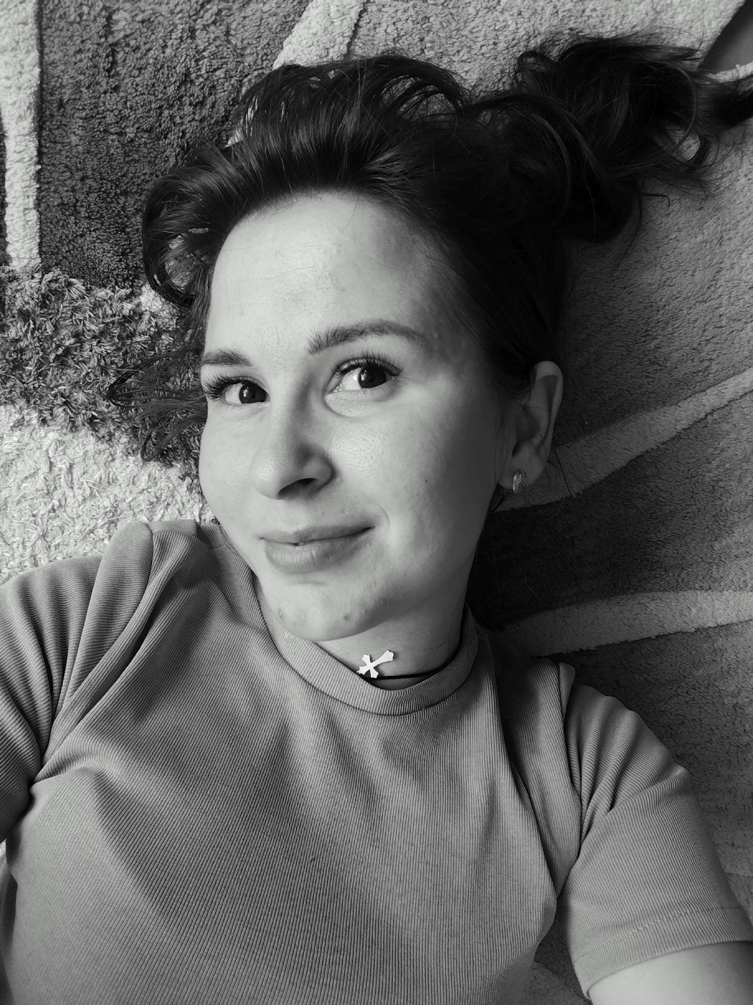 In this black and white, overhead, close-up shot, a young woman with dark hair, pulled up loosely, lies on a textured rug. Her head is tilted slightly, and she looks directly at the camera with a gentle smile. Her eyes are wide, and she appears to be wearing subtle eye makeup. A small, cross-shaped pendant is visible on a choker around her neck. She is wearing a ribbed, short-sleeved shirt. The rug beneath her has a striped pattern, with areas of light and dark texture. The overall impression is one of casual relaxation. The location is identified as Foky, Russia.