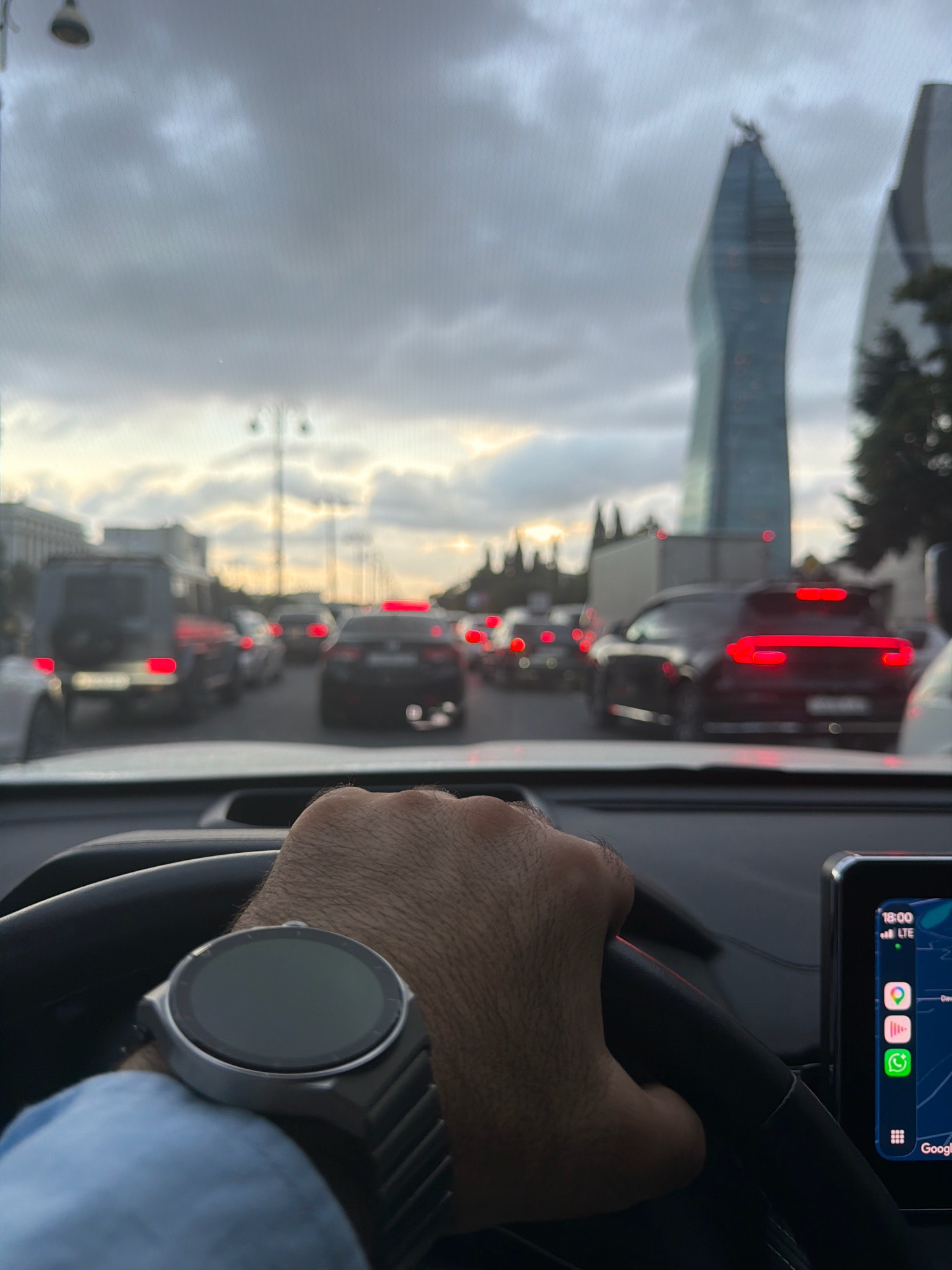 The image is taken from the perspective of a driver in a car, looking forward through the windshield. It is a cloudy day, and the sky is overcast. In the distance, there are tall modern buildings, characteristic of the city skyline of Baku, Azerbaijan, specifically the Flame Towers. The car is in heavy traffic, with multiple vehicles ahead. The driver's left hand, wearing a smartwatch, is visible on the steering wheel. A smartphone is mounted on the dashboard to the right, displaying a navigation app and other icons. The overall scene depicts urban traffic in Baku during daylight.
