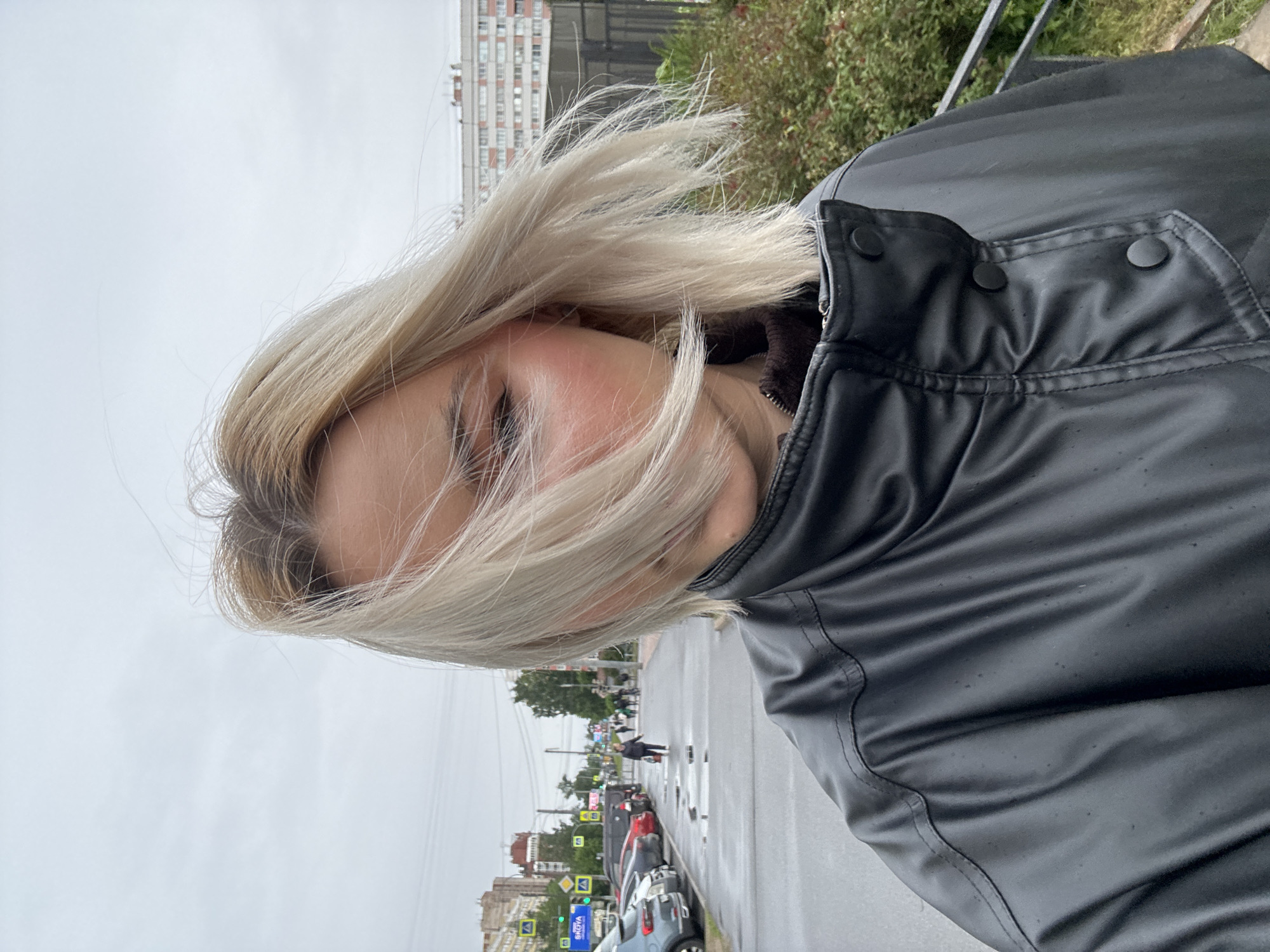 A selfie shot of a fair-skinned woman with blonde, windswept hair standing on a wet street in what appears to be Saint Petersburg, Russia. She is wearing a black, shiny, buttoned jacket. The background features buildings and streetscape typical of a city. A pedestrian is visible crossing the street at a crosswalk. Cars are parked along the street. The sky is overcast.