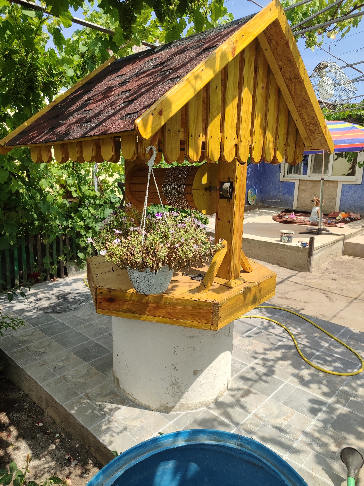 In an outdoor courtyard, a rustic wooden well structure stands in the center, topped with a brown shingled roof. A pot of flowering plants hangs from a hook on the side of the well, and a yellow garden hose coils on the tiled ground nearby. In the background, a blue wall forms the side of a building, with a person sitting on a mat in front of an open window. Green vines with grapes hang from an overhead trellis, and the ground is covered with light-colored tiles.