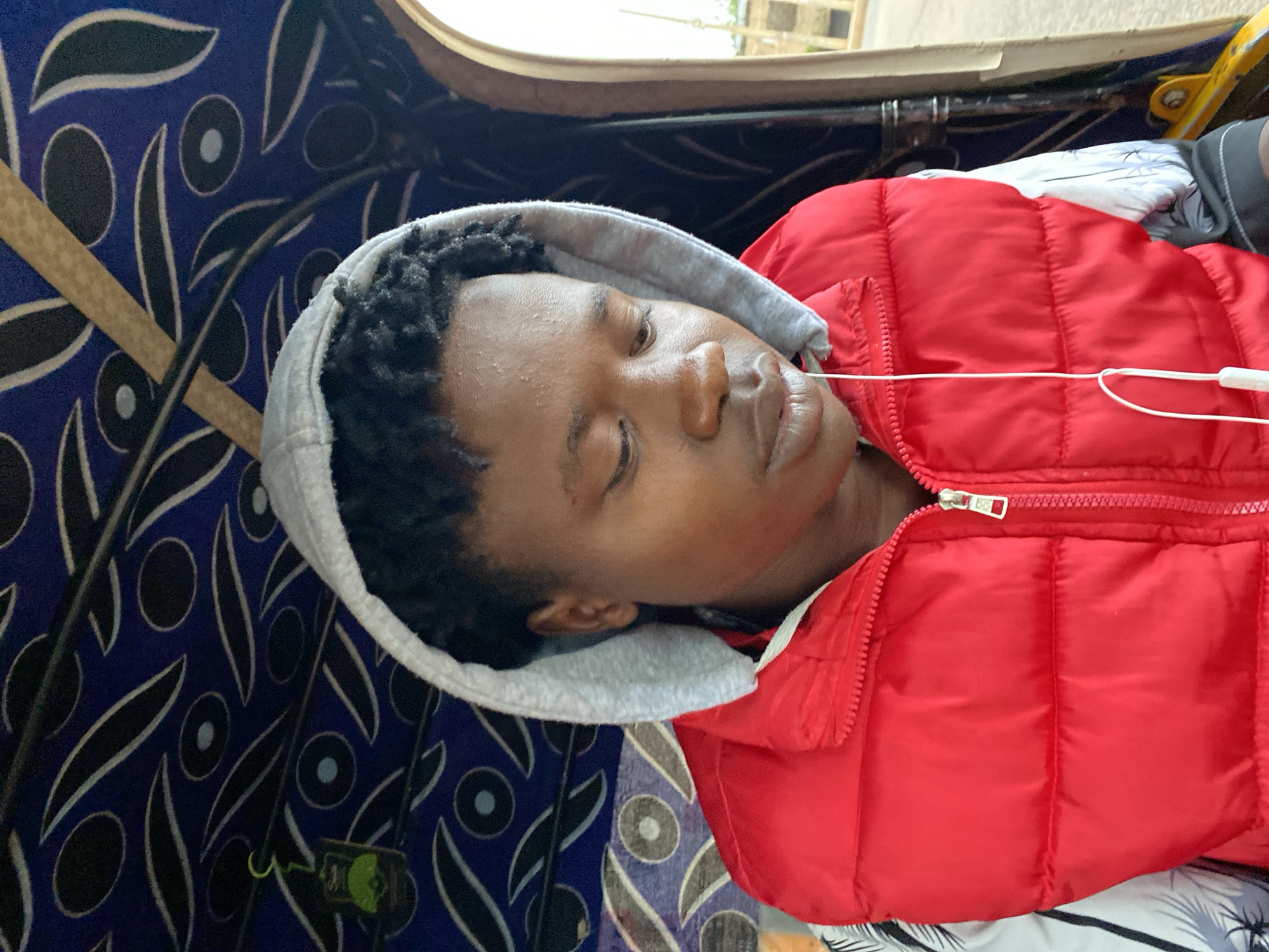 A young Black male with dark, curly hair and a light grey hooded sweatshirt pulled over his head is seen from the chest up, looking downwards and to his right. He is wearing a bright red puffer vest zipped up to his neck, over a white shirt with a black and white patterned design. A white earbud wire hangs from his ear and rests on his vest. The background appears to be the interior of a vehicle, possibly a tuk-tuk, with a dark blue canopy featuring an abstract pattern of black and white shapes, including ovals and eye-like motifs. A light green tag with a black silhouette is visible hanging from a hook on the left side of the canopy. The overall lighting suggests an outdoor setting.