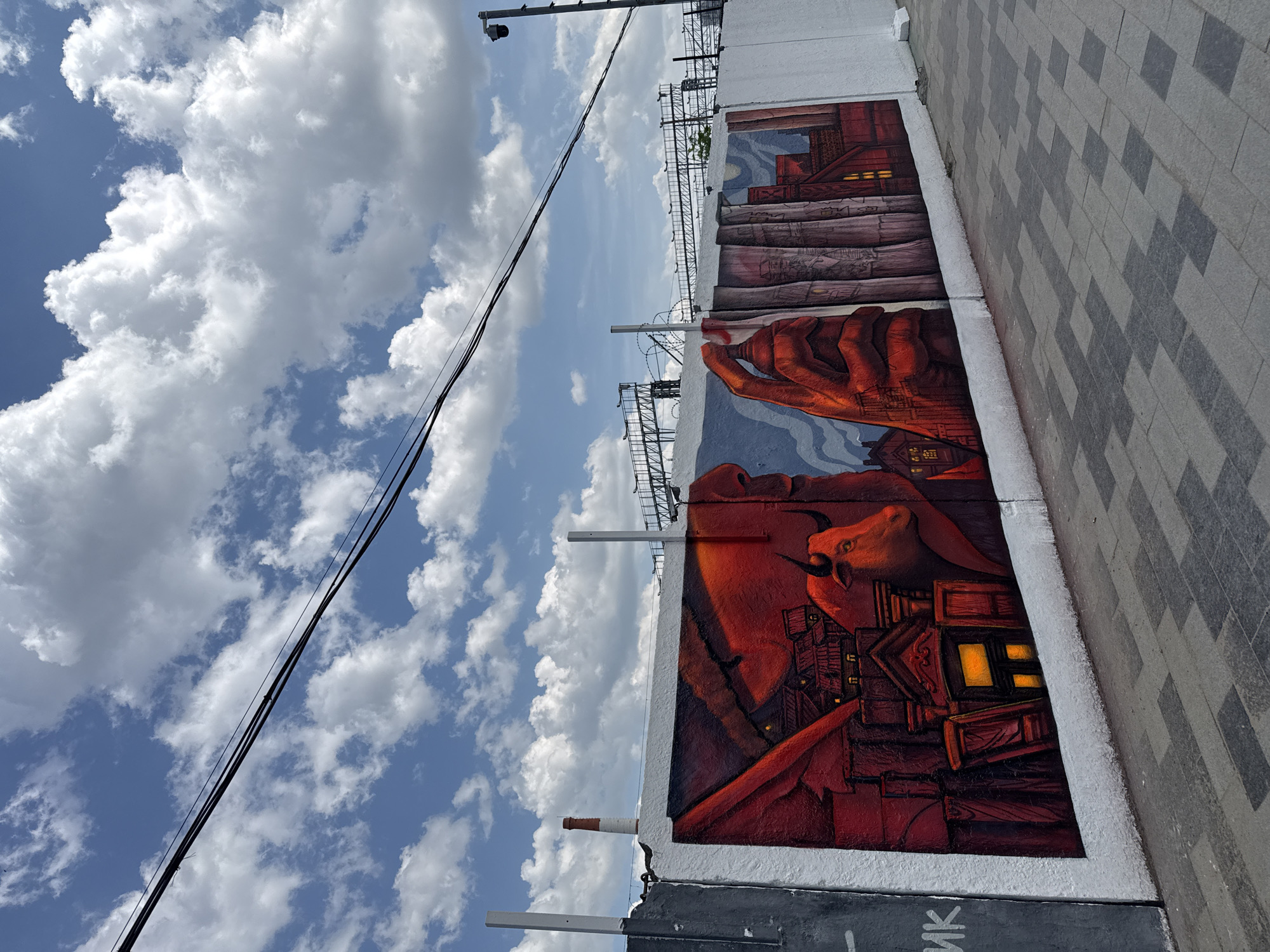 A mural adorns a wall adjacent to a pedestrian walkway, likely in Moscow, Russia, given the Cyrillic characters visible. The mural depicts a bull, a hand holding what appears to be a building, and cityscapes, all rendered in shades of red, orange, and blue. The sky above is blue with puffy white clouds, and electrical lines run across the upper-left of the frame. A security camera is mounted on a pole above the wall. The walkway is paved with gray and white rectangular blocks.