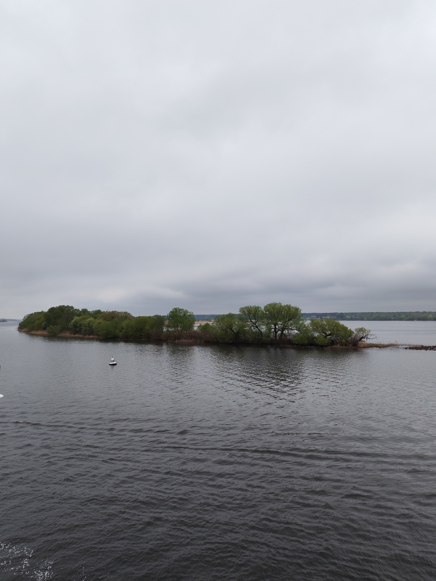 The image depicts an island covered in trees in the middle of a body of water, likely a river or lake. The sky is overcast. A white buoy is visible in the water near the island. In the background, a distant shoreline with trees can be seen. The water is dark and rippled. The location is not explicitly stated in the image or provided in the given context. Therefore, it is not possible to confirm that the location is Sloboda, Russia without additional information.