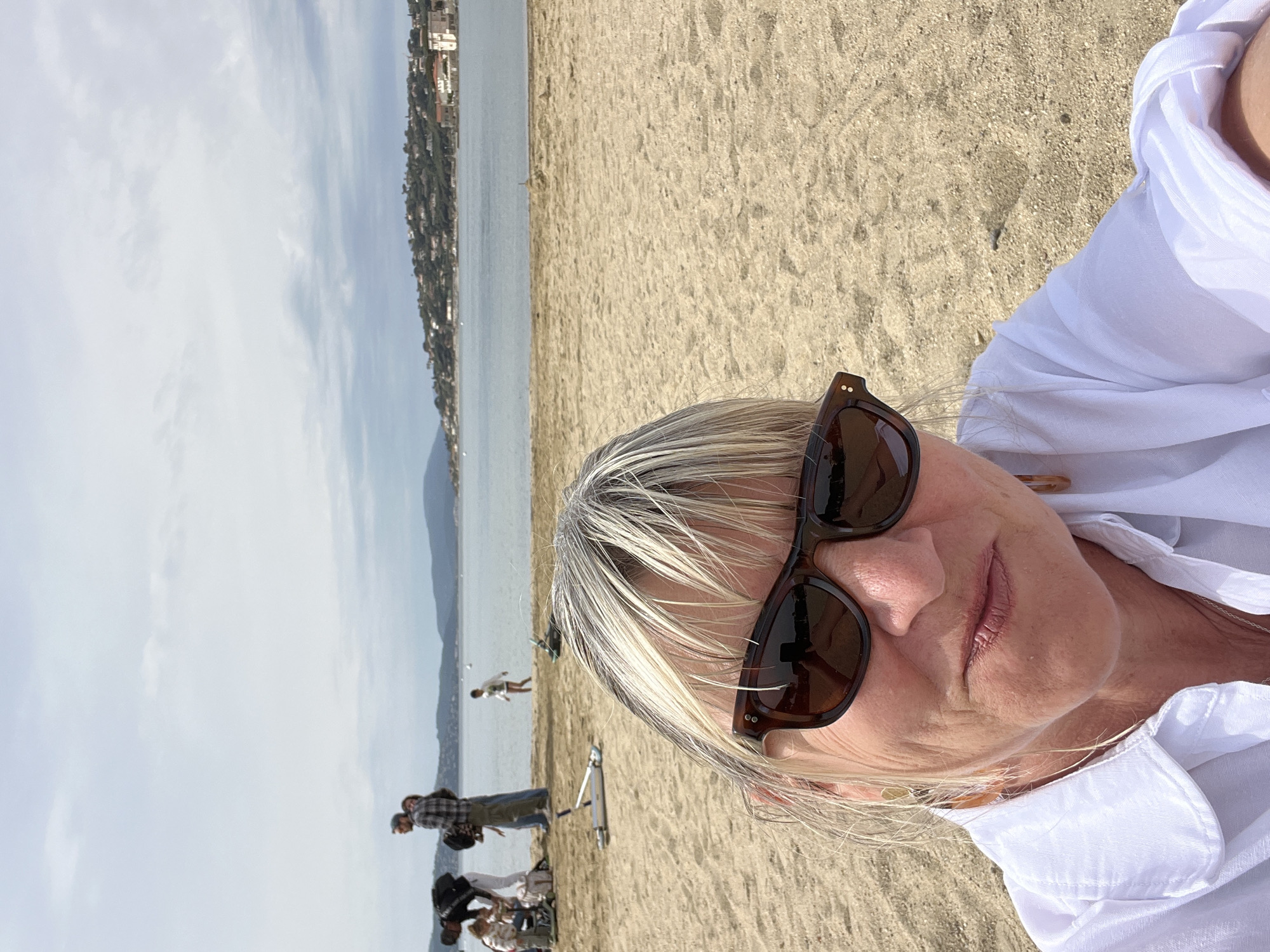 A medium-shot selfie features an older woman with gray and blonde hair, wearing sunglasses and a white shirt. The background shows a beach with sandy ground, a body of water (likely the sea), and a distant cityscape along the coastline, indicating the location. Several people are visible on the beach, including a child running, and groups of people standing and interacting. The sky is pale and slightly overcast. Given the query context, the location is likely a beach in Sainte-Maxime, France.