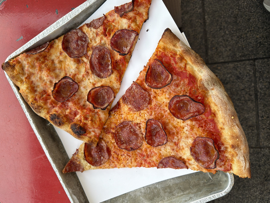 Two triangular slices of pepperoni pizza are presented on a sheet of white paper, which rests inside a rectangular metallic tray with raised edges. The pizza slices feature a red tomato sauce base, melted white cheese, and numerous circular pepperoni slices, some of which are curled and exhibit darkened edges. The crusts are golden-brown with some charred areas. The metallic tray is positioned on a worn, reddish-orange tabletop. A dark, textured surface is visible in the bottom-right background, beyond the table. The scene is static. This scene is located in the city of Tel-Aviv, Israel.