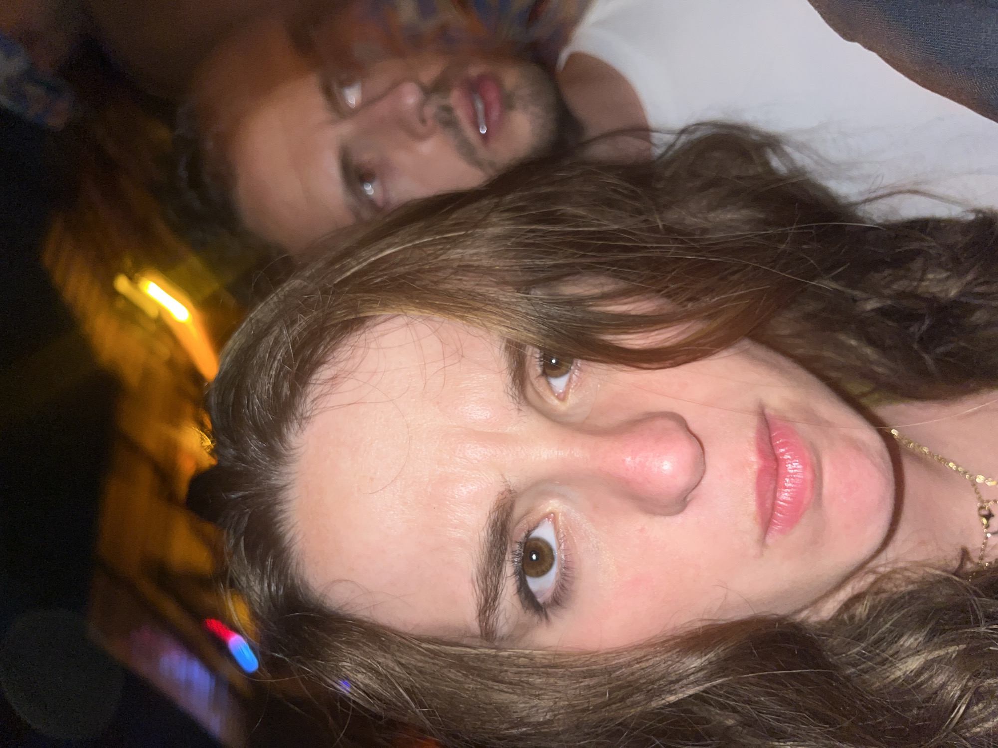 A medium-angle shot depicts two people in Puerto Escondido, Mexico. The woman is in the foreground, her face prominent. She has light skin, brown eyes, and brown hair. Her lips appear to have pink lipstick. She is wearing a delicate gold necklace. Behind her is a man with light skin, brown eyes, and dark hair. He wears a white t-shirt. There are blurry lights and colors in the background, possibly from a nightlife scene.