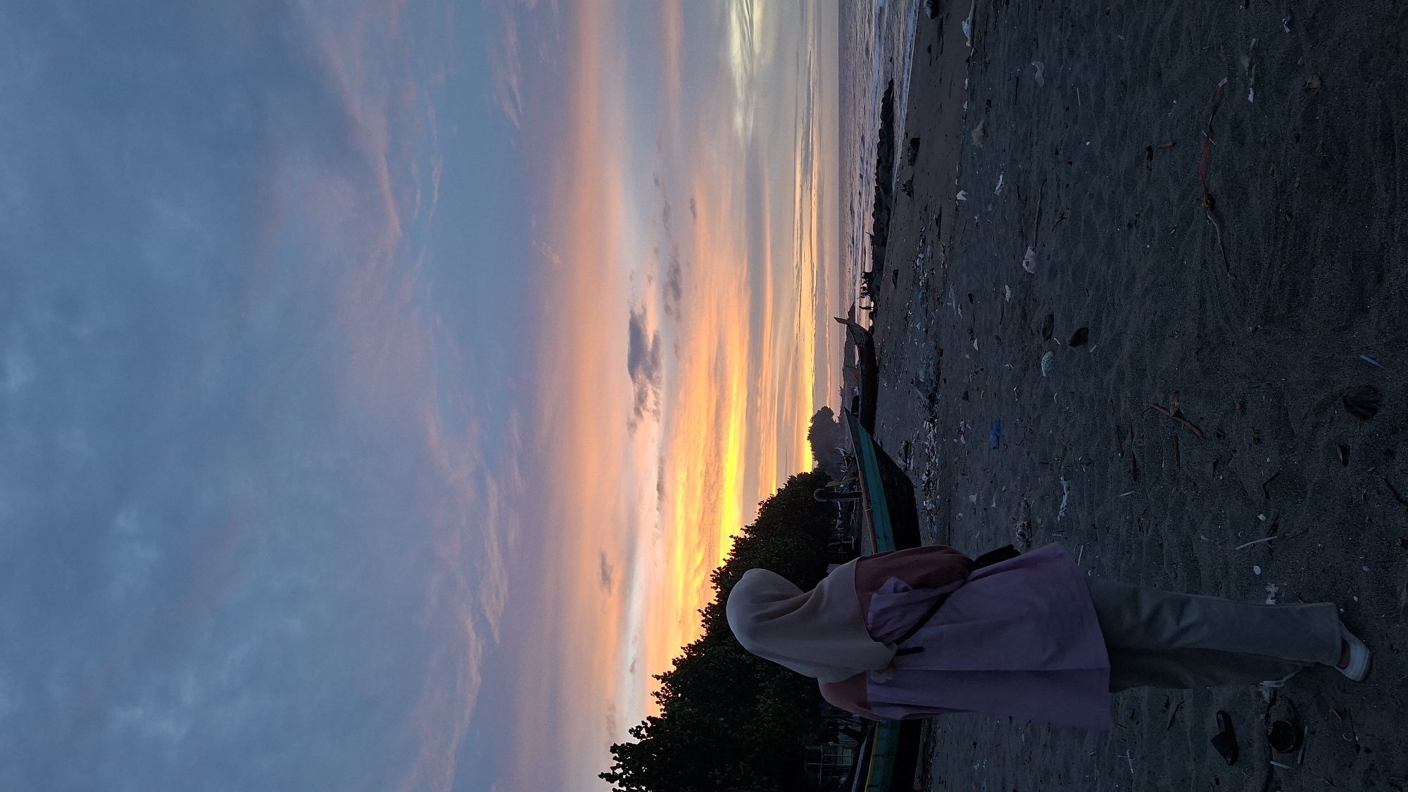 A person wearing a light-colored hijab and a pink and light brown top stands on a dark sandy beach, facing away from the camera towards the ocean and a vibrant sunset. The sky is filled with streaks of orange, yellow, and pink clouds, transitioning to blue at the top. Several fishing boats are visible on the beach, some partially submerged in sand. The shoreline is littered with debris, including plastic waste. In the background, a line of dark green trees separates the beach from the sky. The overall scene depicts a serene yet somewhat unkempt coastal environment at dusk.