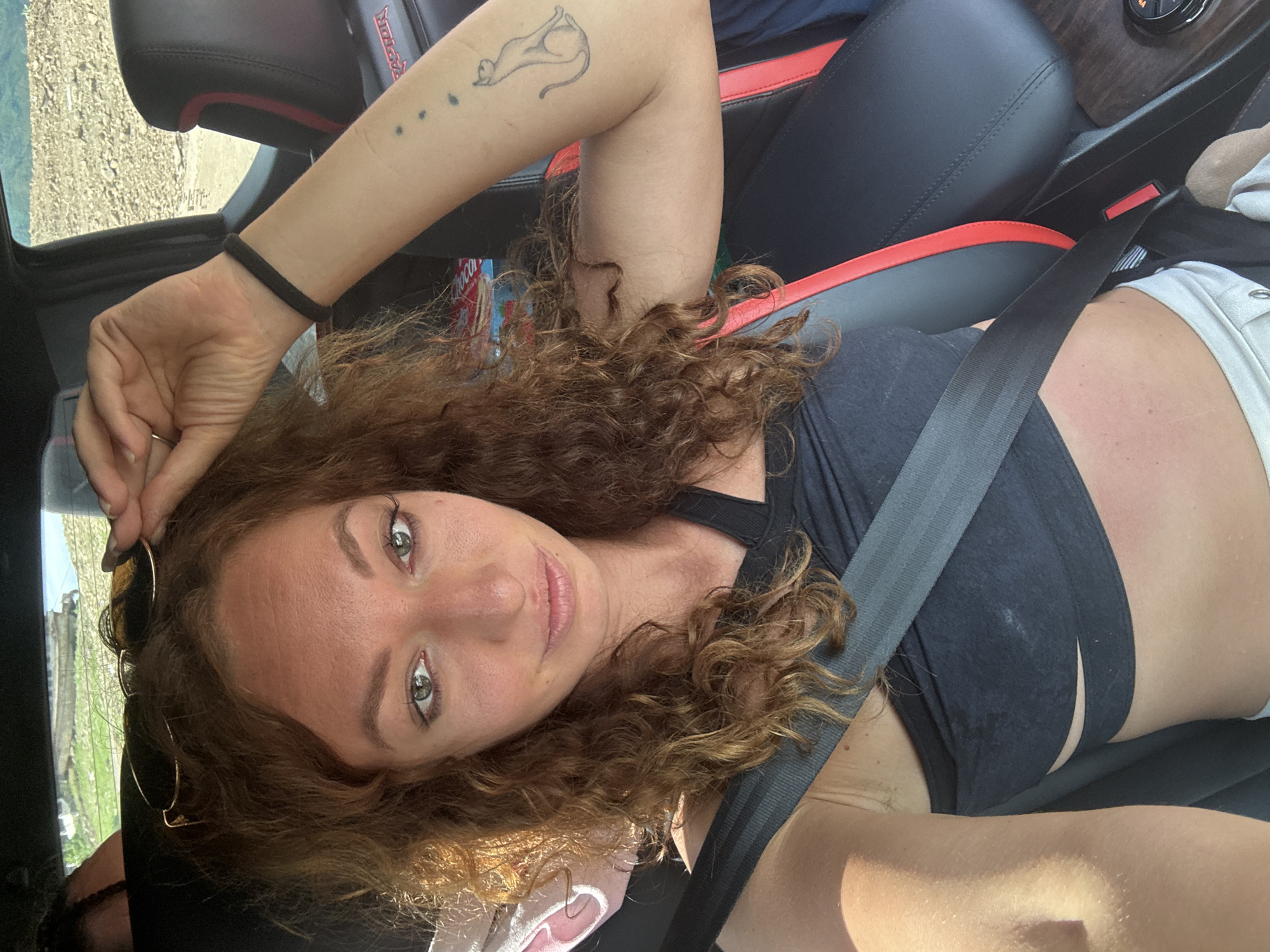 A woman with curly brown hair is in the driver's seat of a car. She is wearing a black crop top and white shorts, and a seatbelt is across her chest. She is holding a pair of sunglasses above her head. Her left arm, which has a tattoo of a cat and paw prints, is resting on the armrest of the car seat. The car's interior is black with red trim. The background shows a rocky, outdoor landscape. The image does not contain information related to Tân Phong, Vietnam.