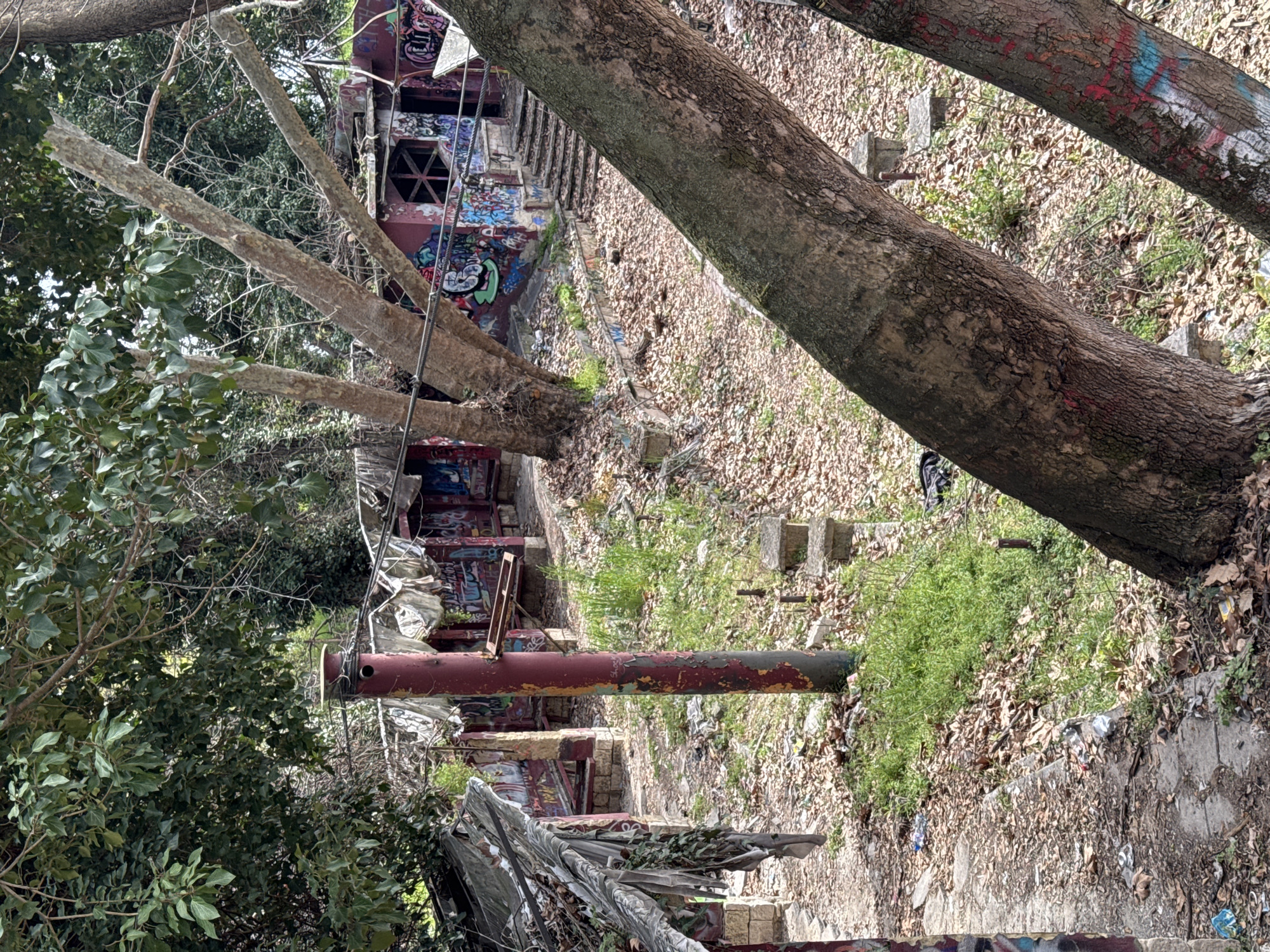 The image depicts a decaying, graffiti-covered structure in what appears to be an overgrown area. The structure is multi-tiered, with concrete steps and walls heavily marked with graffiti. A large metal pipe is visible, and surrounding foliage suggests an abandoned location. The presence of trash and debris indicates neglect. The angle of the shot and surrounding vegetation obscure a full view of the site. Given the distinctive graffiti style and the type of construction, this could be the abandoned Old Baths of Varna, Bulgaria.