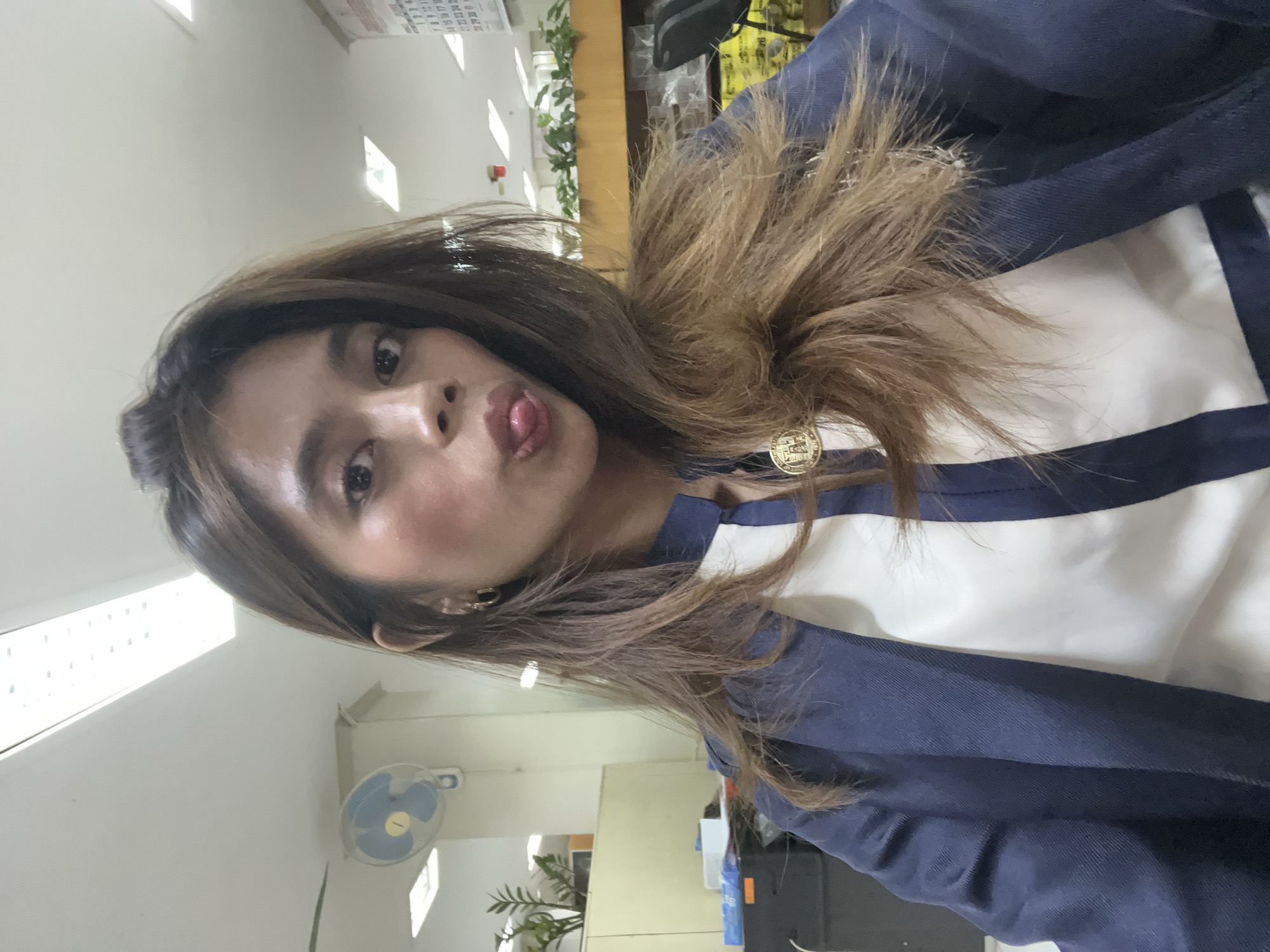 A woman with long, brown hair is taking a selfie in what appears to be an office environment. She is wearing a dark blue blazer over a white top with a dark blue stripe. She is sticking her tongue out and making a pouty face. In the background, there are fluorescent lights on the ceiling, a wall-mounted fan, plants on a shelf, and a calendar on the wall. The presence of office equipment and supplies suggests the location is likely a business or administrative office in Davao City, Philippines.