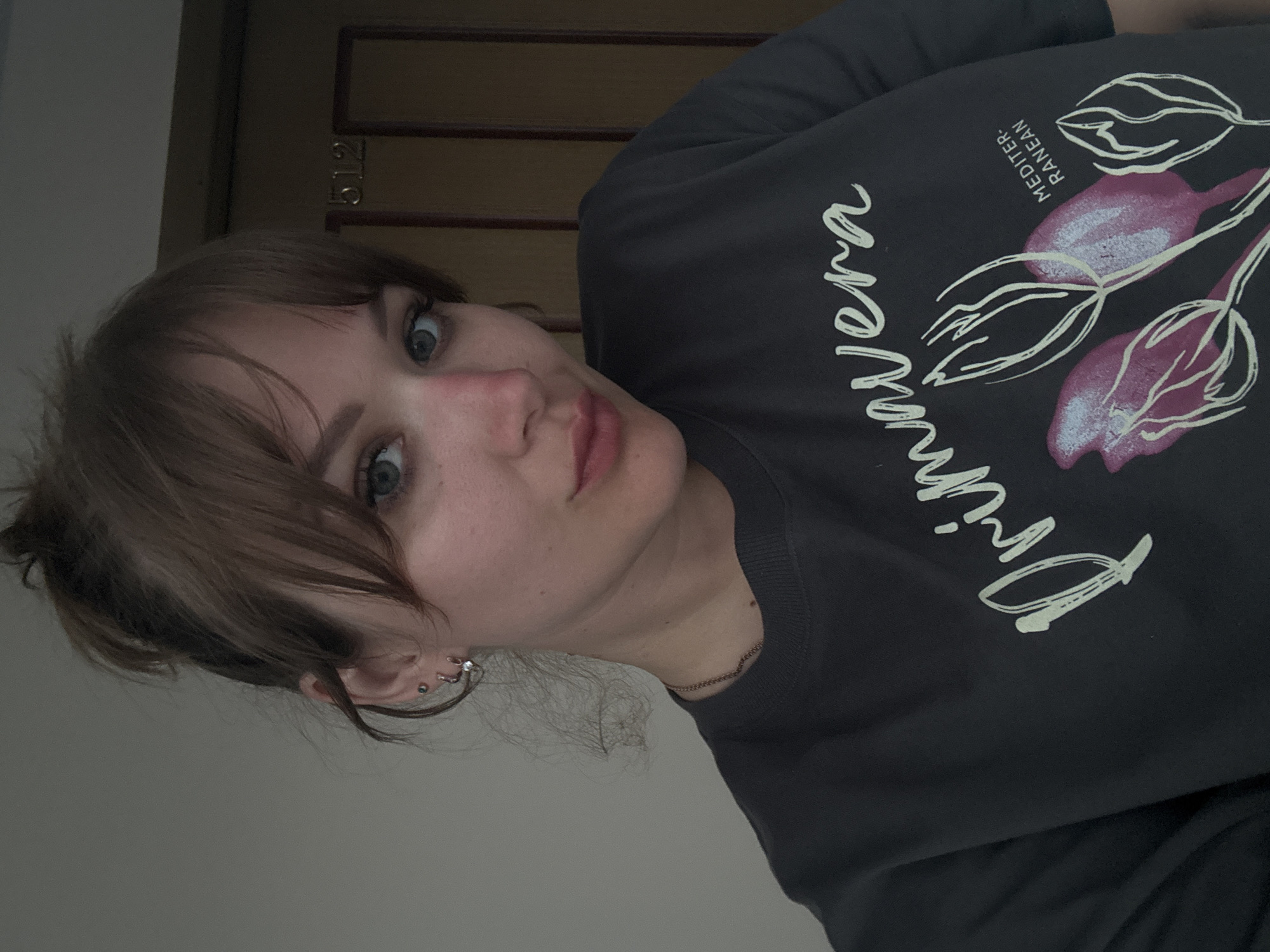 A young woman with fair skin and light brown hair is posing indoors. She has blue eyes and is wearing a dark grey shirt with a white floral design and the word "Primavera" written in cursive. She has multiple earrings in her left ear. The background includes a light-colored wall and a dark brown door with a gold-colored doorknob. The lighting appears to be artificial. The location in the city of Yevpatoriya, Ukraine, cannot be determined from this image.