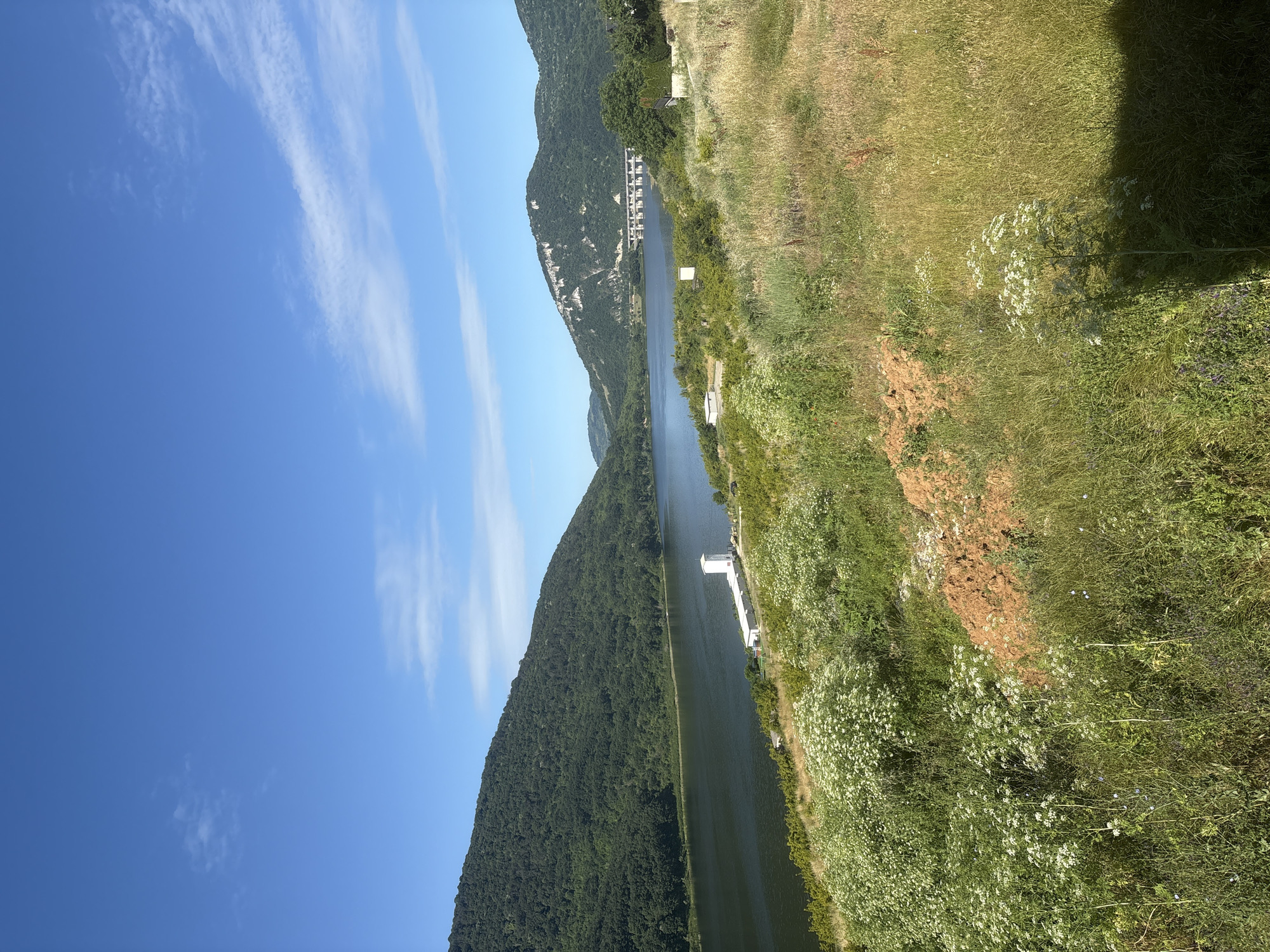 A landscape scene captures a wide, calm body of water, likely a river or lake, flanked by lush green, tree-covered mountains on one side and a grassy slope dotted with wildflowers on the other. The water reflects the blue sky and the surrounding landscape, creating a sense of depth and tranquility. On the far side of the water, a white structure, possibly a pier or a boat dock, is visible. Further along the water, a series of buildings or structures are nestled into the mountainside, suggesting a small settlement or resort. The sky is a vibrant blue with wisps of white clouds, indicating a clear, sunny day. The time of day appears to be late morning or early afternoon, based on the bright sunlight and the angle of shadows. The overall mood of the image is peaceful and serene, evoking a sense of natural beauty and escape. There is no visible text in the image.