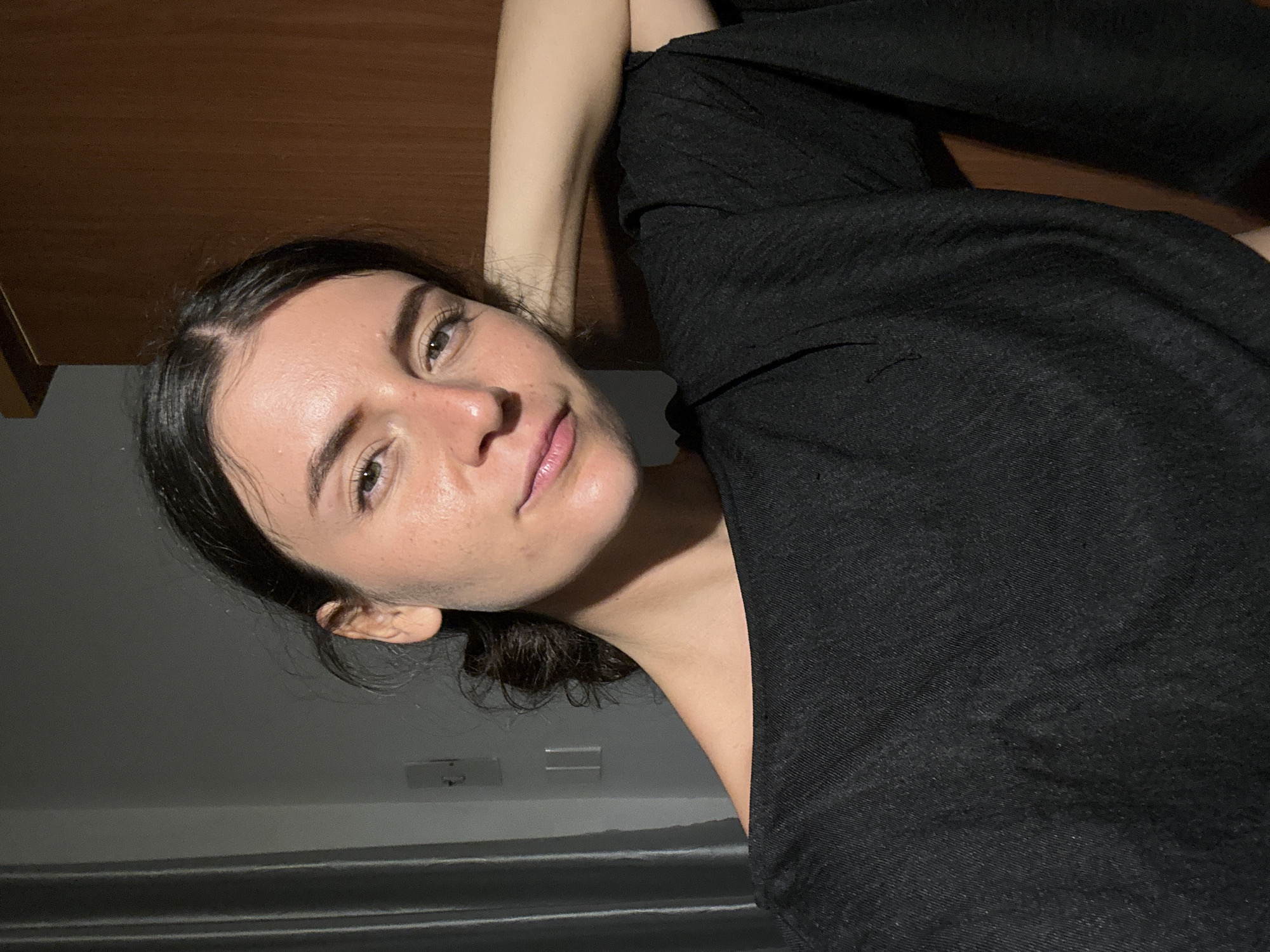 A woman with dark hair and fair skin is lying down, looking at the camera. She is wearing a dark-colored shirt. The background consists of a wooden surface above her and a gray wall with a possible electrical outlet below her. The image does not provide sufficient information to determine a specific location in Mũi Né, Vietnam.
