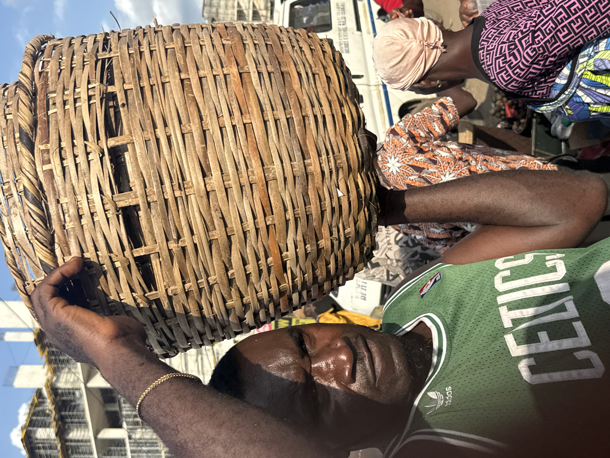 In Owerri, Nigeria, a Black man, visibly sweating, carries a large, woven basket on his head. He is wearing a green Adidas Celtics basketball jersey. His left arm, adorned with a gold bracelet, is raised to help balance the basket. Behind him, other individuals are visible, including a woman wearing a pink headscarf and a patterned top, and another person in an orange and white patterned shirt. In the background, a white vehicle with markings, possibly a delivery truck, is parked, and construction is underway on a building with scaffolding. The scene appears to be a busy outdoor market on a bright, sunny day.