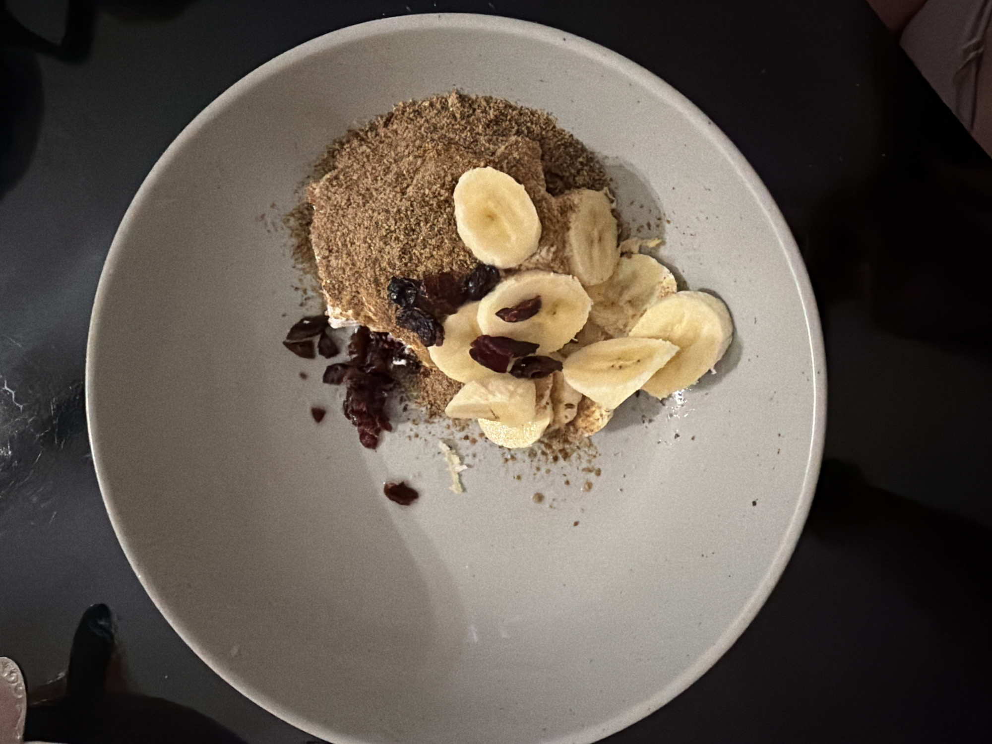 A top-down view shows a light grey bowl filled with a breakfast meal, resting on a dark surface. The meal consists of what appears to be a mound of granola or ground flaxseed, sliced bananas, and dark dried cranberries or raisins. No people or specific environmental details relating to Pervomaysky, Russia are visible.