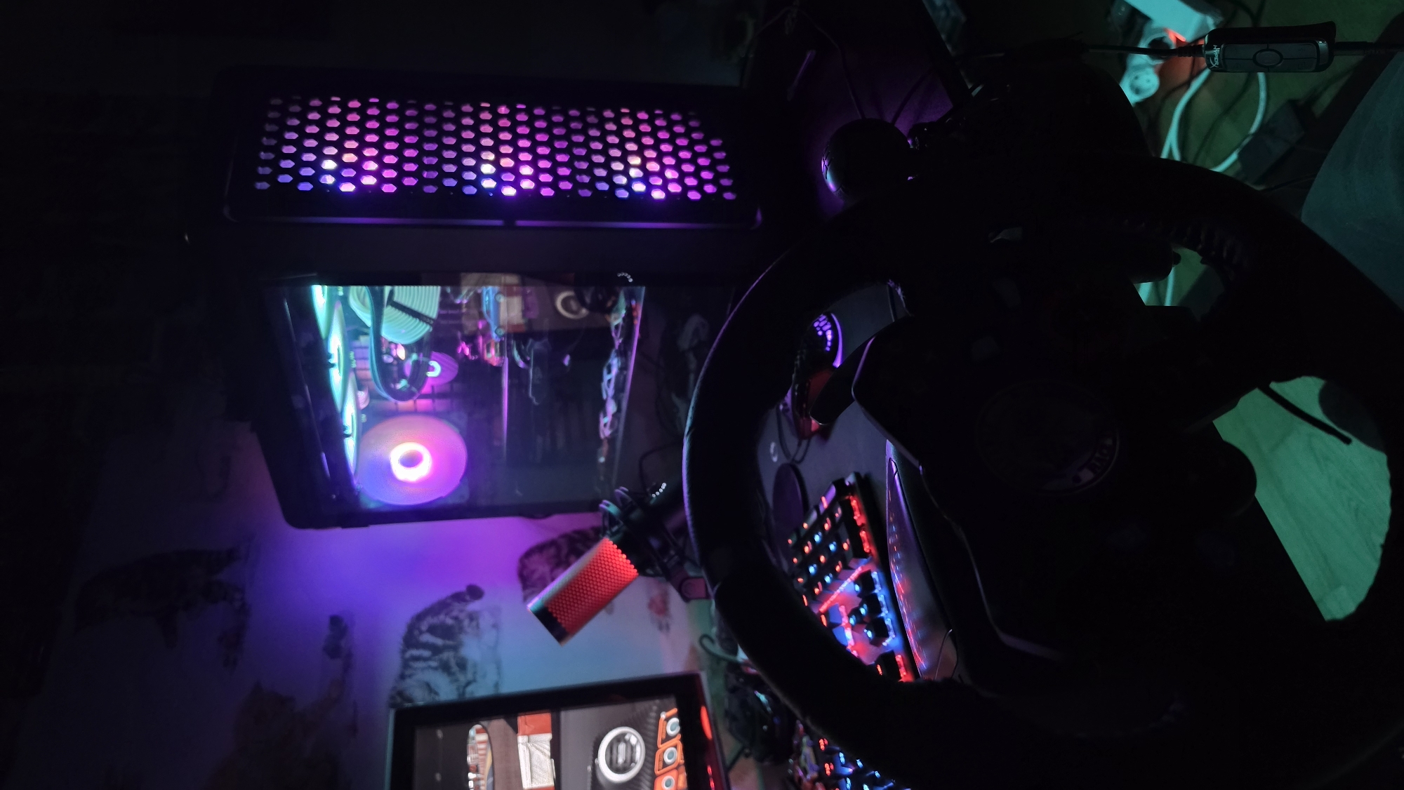 The image captures a dimly lit, indoor gaming setup, likely at night, characterized by vibrant RGB lighting. The central focus is a gaming PC tower on the left, featuring a mesh top panel illuminated with pink and purple lights. Inside the PC case, multiple fans emit purple and teal glow, with visible liquid cooling tubes.
Dominating the right side of the frame is a large, dark gaming steering wheel, its form silhouetted against the ambient light. A circular emblem or logo is visible on its center. Beneath the steering wheel, a mechanical keyboard with red and blue backlighting is partially visible. A bright red, cylindrical microphone is mounted on an arm, positioned near the lower center.
A section of a monitor screen is visible at the bottom of the image, displaying graphical elements that appear to be part of a game interface or menu with circular designs. The diverse RGB lights from the PC and peripherals cast colorful ambient light, primarily purple, teal, and green, onto the surrounding walls and objects. Faint, abstract patterns are discernible on the wall behind the setup, illuminated by the colorful glow. Various wires and cables are also present, typical of a complex electronic workstation. No people are visible in the scene. The specific location within Varna, Bulgaria, is not identifiable from the image.