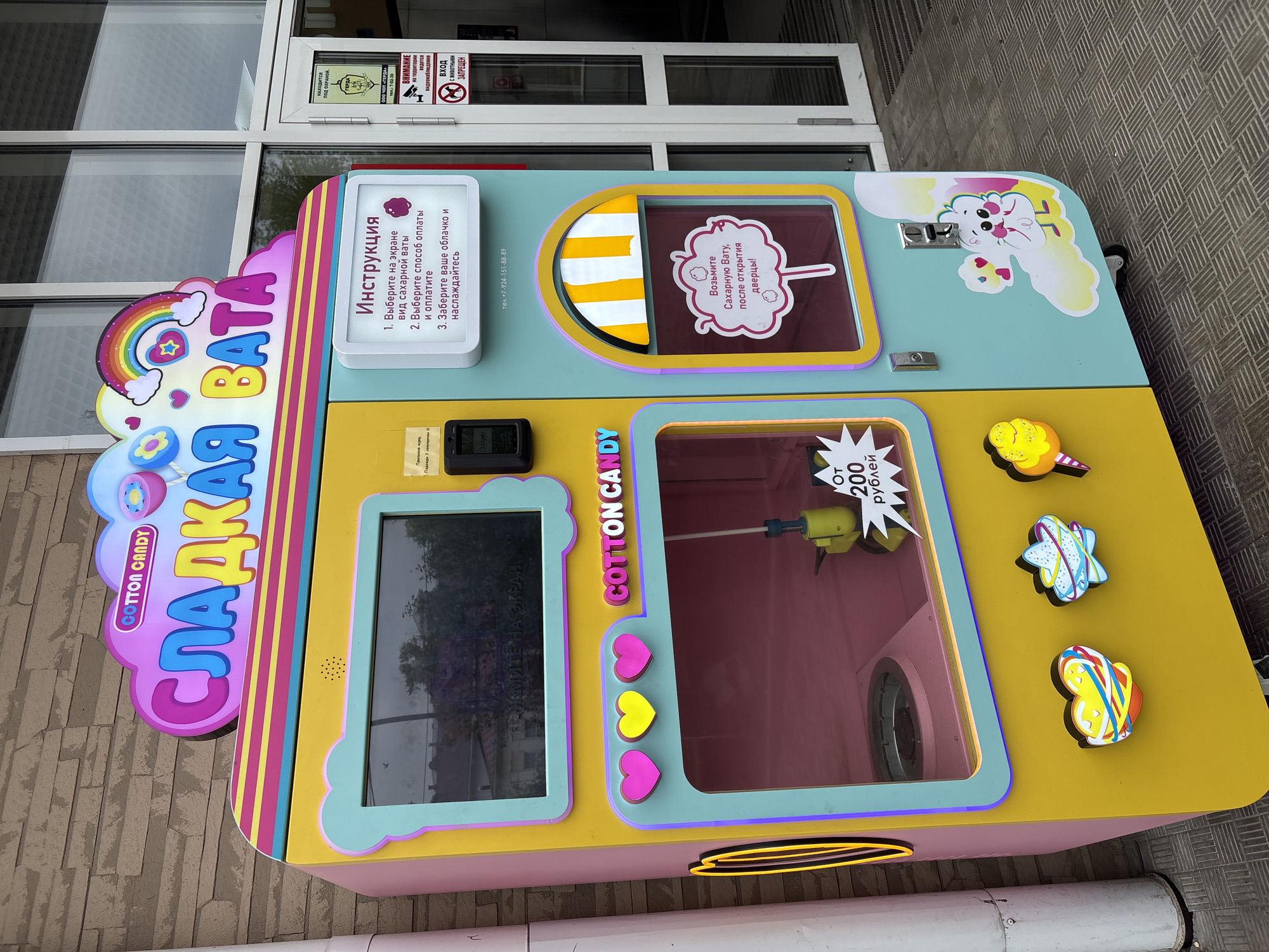 The image shows a close-up, high-angle shot of a cotton candy vending machine located in Birobidzhan, Russia. The machine is brightly colored, with pink, yellow, and blue hues, and features cartoonish illustrations. A large sign on the left side spells out "СЛАДКАЯ ВАТА" in Cyrillic, meaning "Sweet Cotton," with "COTTON CANDY" written in English underneath. The machine includes a screen, presumably for ordering and payment, and a window to view the cotton candy being made. There are instructions in Russian on how to use the machine. The machine includes signage stating the cost, starting from 200 rubles. The background includes part of a building facade with a window and other signs, including a notice that the area is under video surveillance and that animals are not permitted. The ground appears to be paved with a patterned surface.