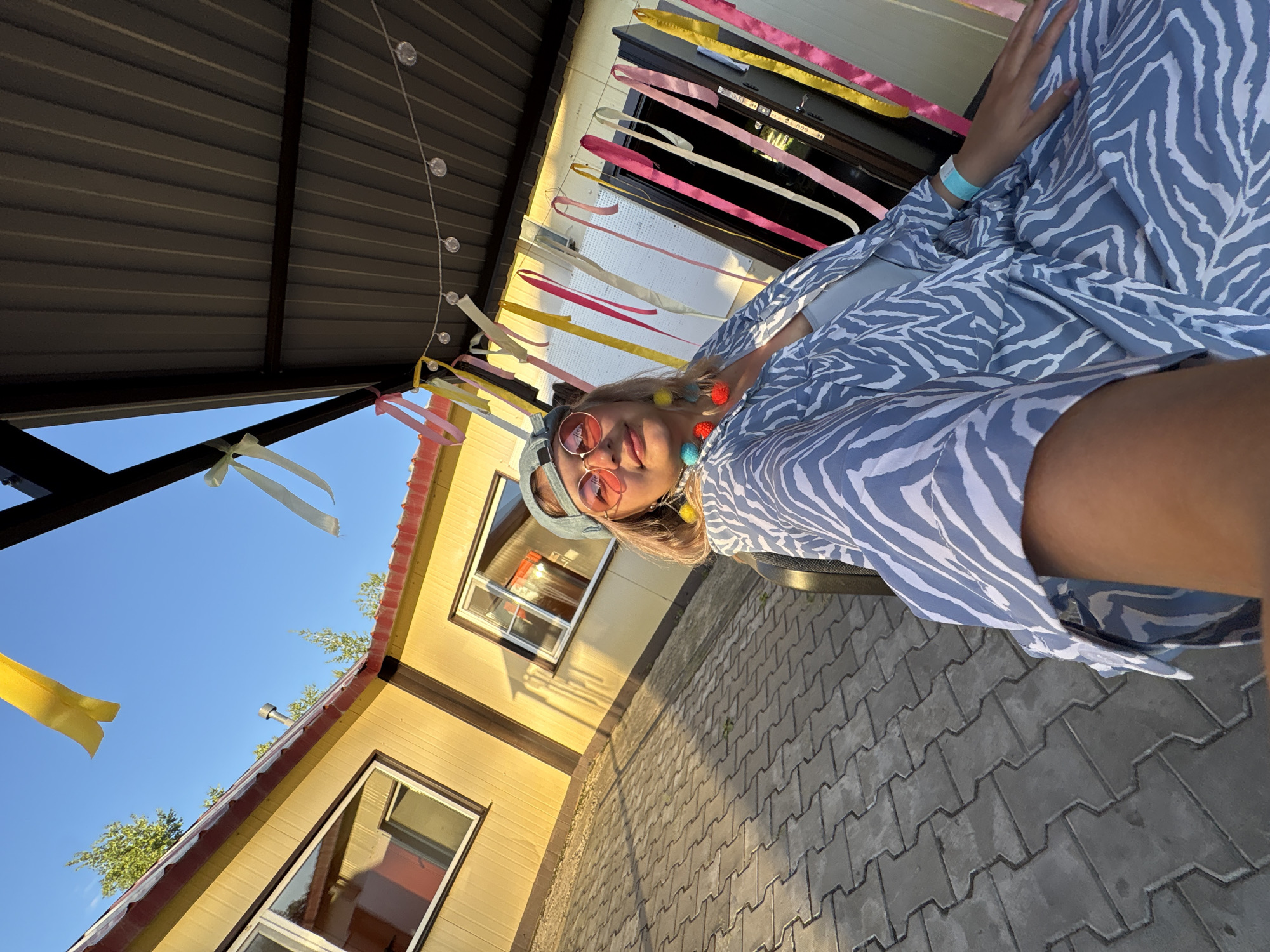 A young woman is posing for a selfie in front of a yellow building with a red-trimmed roof in Birobidzhan, Russia. She is wearing a blue and white zebra-print robe, a teal baseball cap, and pink-tinted sunglasses. Colorful ribbons and string lights are draped from the overhang of a dark-colored roof structure behind her, creating a festive atmosphere. The ground is paved with gray interlocking stones. The sun is casting long shadows, indicating it is either early morning or late afternoon. The sky is clear and blue.