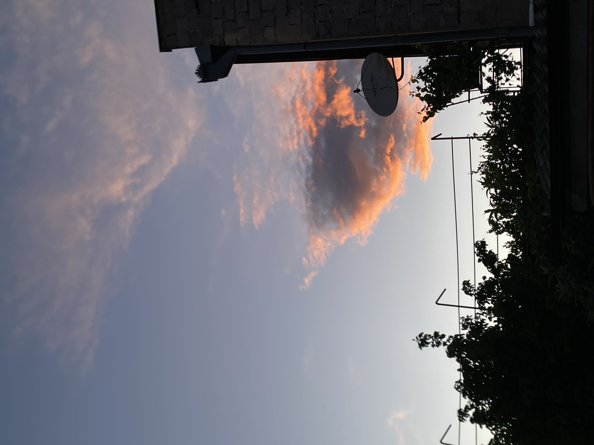 The image, rotated for viewing, shows a dusk sky with orange-tinged clouds near a building. A satellite dish is mounted on the building's edge. Vines or plants are visible on a balcony or structure to the right, silhouetted against the sky. A section of brick or stone wall is seen at the top. The location is likely Tbilisi, Georgia.