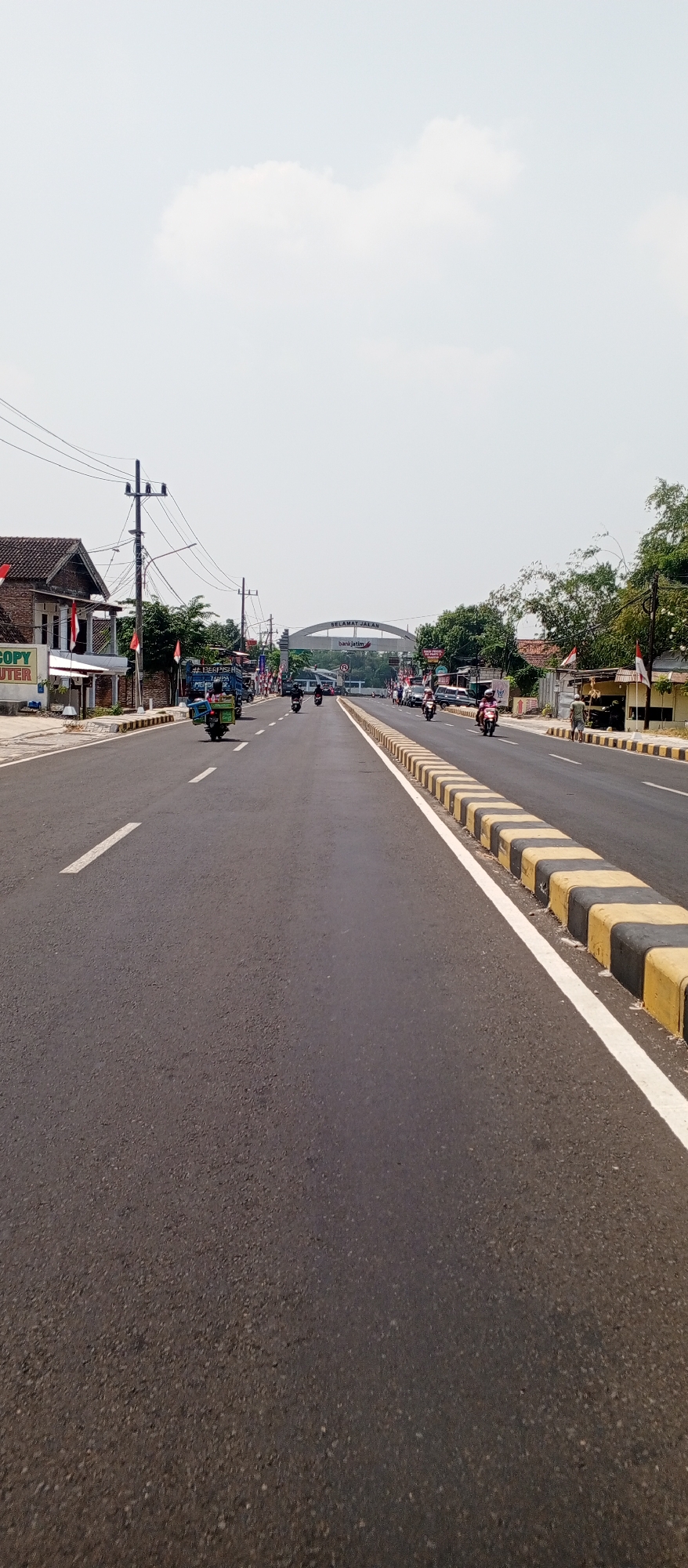 This is a vertical, eye-level, medium-shot photograph taken on a sunny day in Maospati, Indonesia. The scene depicts a two-lane asphalt road leading towards an arched gateway. The gateway has the text "SELAMAT JALAN" and "bankjatim" written on it. The road is marked with a white dashed line down the center and a white solid line on the right side. The right side of the road is bordered by yellow and black striped concrete barriers.
On the left side of the road, there are buildings and a power pole. One building has a sign that reads "COPY CENTER". Red and white Indonesian flags are visible on poles along the roadside. Several motorcycles and a blue truck are driving on the road. Some people are riding the motorcycles. Trees and other vegetation line the sides of the road, and some houses are visible in the background. The sky is clear with a few scattered white clouds.