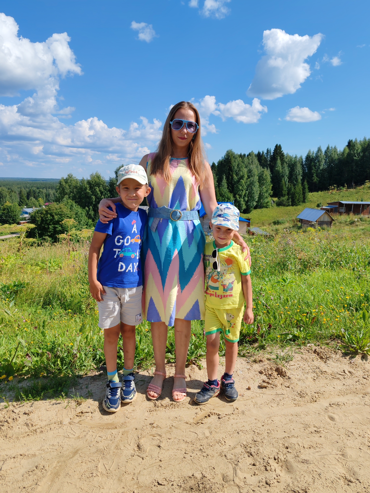 In the city of Chukhloma, Russia, under a bright blue sky with scattered white clouds, a woman stands with two young boys on a sandy path. The woman, wearing a multi-colored geometric dress and blue sunglasses, has her arms around the boys. The boy on the left wears a blue t-shirt with a graphic print, light-colored shorts, and a white baseball cap. The boy on the right is dressed in a yellow t-shirt and shorts with a cartoon print, and a blue and white camouflage baseball cap. Behind them, a lush green landscape unfolds, featuring a dense forest of evergreen trees. In the distance, several wooden houses are visible, suggesting a rural or semi-rural setting. The foreground is dominated by the sandy path and the vibrant green grass and wildflowers.