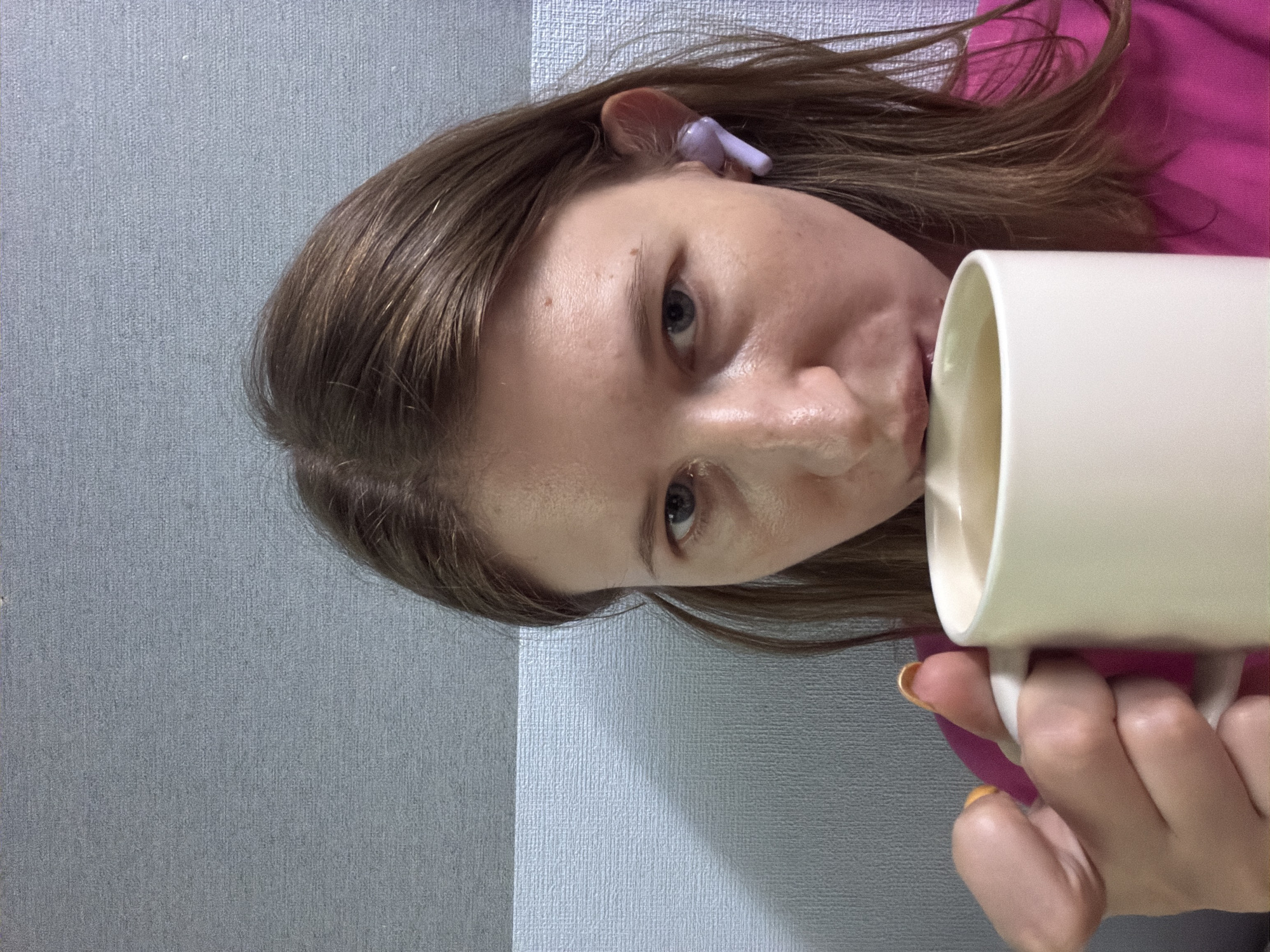 A medium close-up shot shows a woman holding a white mug to her lips, as if drinking. She has long, brown hair, fair skin, and is wearing a bright pink shirt and a purple wireless earbud in her right ear. The background appears to be a textured wall with horizontal bands of light and dark grey. The image does not provide any specific information to determine its location within Dongducheon-si, South Korea.