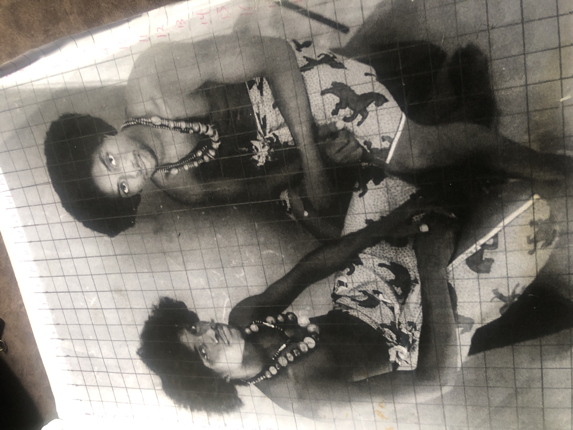 The image displays a black and white photograph of two men, seemingly posed. Both men have dark, curly hair and are wearing necklaces with large, circular beads. They are also dressed in patterned wraps or skirts. The photograph is overlaid with a grid, suggesting it might be a reference image for a drawing or painting. Numbers are visible along the right edge of the grid, ranging from 10 to 16, and possibly continuing further. The background is indistinct, but grid lines suggest a paper surface. There is no discernible information within the image that places it in Ado, Nigeria, or any specific urban location. The individuals' attire does not provide a definitive cultural or geographical marker for Ado, Nigeria, without additional context.