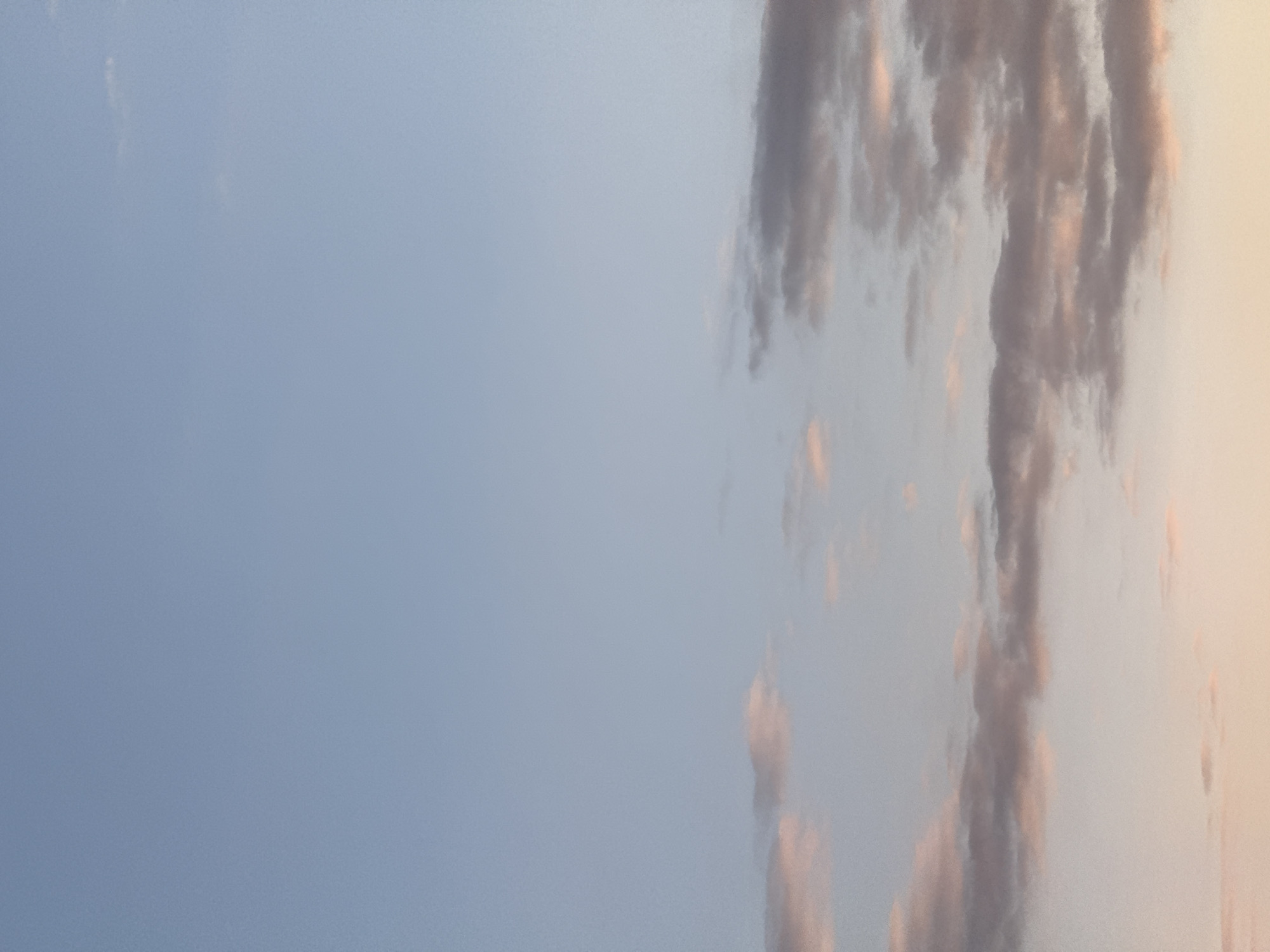 The media file displays a sky scene dominated by a gradient of colors. The left side of the image features a clear, soft blue-grey sky. As the view shifts towards the right, a warmer, peachy-orange hue emerges, indicative of either sunrise or sunset. A substantial formation of clouds occupies the right portion of the frame, appearing as a vertical band. These clouds are textured, varying in shades of grey and purplish-brown, and are illuminated by the warm, peachy light, creating highlights of orange and pink. There are no discernible people, objects, or activities taking place. No text is visible. The scene captures a tranquil atmospheric moment, with the light suggesting either dawn or dusk.