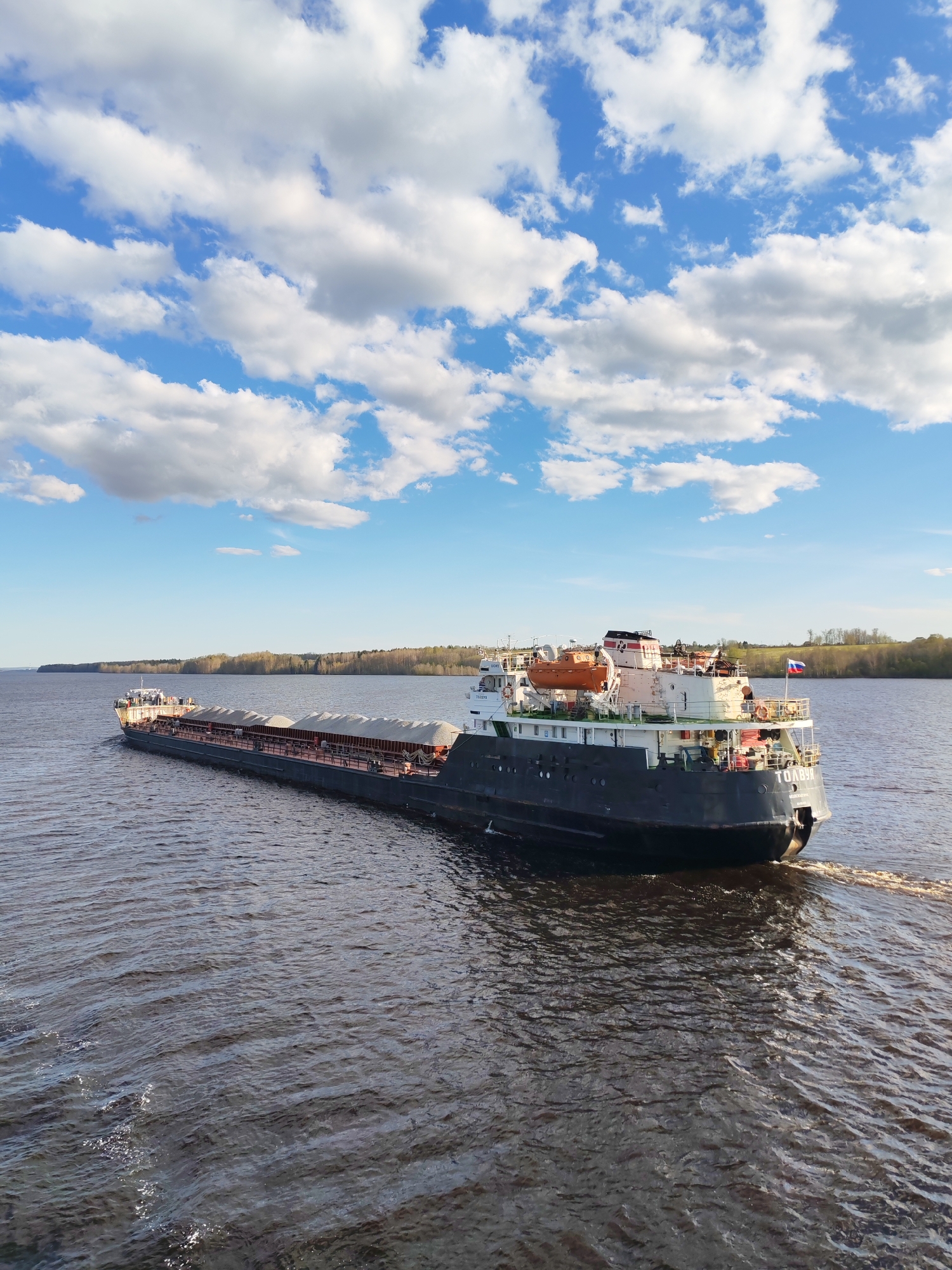 A barge, named "Tolvuya," is on a river in Yakunina Gora, Russia. The barge is black and carries a cargo of gravel or a similar material. The stern of the barge is visible, and it has a Russian flag. The river is brown and slightly rippled. In the background, there is a distant shoreline with trees. The sky is blue with scattered white clouds. The weather appears sunny and calm.
