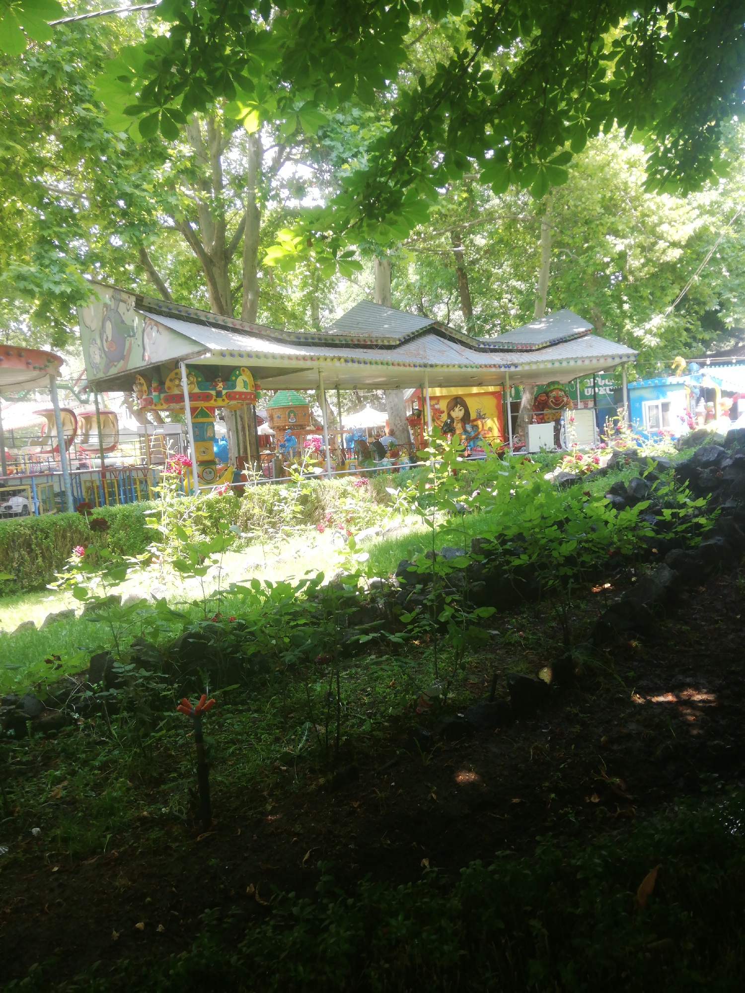 The image shows a colorful carousel ride in a park. The carousel is adorned with cartoon characters and is surrounded by lush green trees and bushes. In the foreground, there are some red flowers and dark rocks. The scene appears to be set in a public park on a sunny day, possibly in Yerevan, Armenia, given the context provided. There are no people clearly visible in the image, but the presence of the carousel suggests a place of recreation. The overall atmosphere is bright and cheerful, with the sunlight filtering through the leaves creating a dappled effect. The carousel itself seems to be in good condition, with vibrant colors and intricate details on the ride vehicles and supporting structures.
