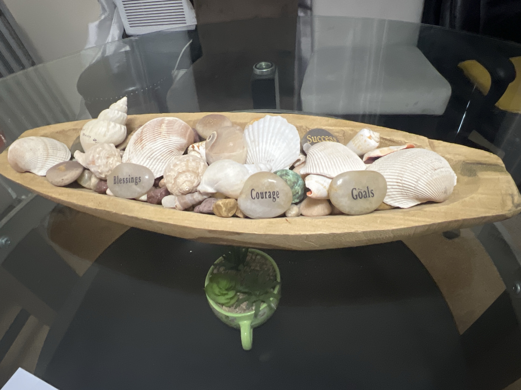 The image shows a wooden bowl filled with seashells and stones inscribed with the words "Blessings," "Courage," "Success," and "Goals." The bowl is placed on a glass-topped table. Underneath the bowl and on the table sits a small green cup containing succulents. The background is blurry, revealing a glimpse of a white object. The location cannot be determined from the image. There is no discernible evidence to suggest that this photo was taken in Chicago, United States of America. There are no people visible in the image.