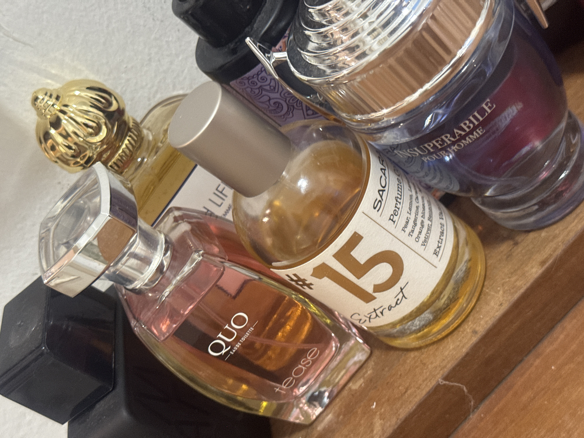 The image shows a collection of perfume bottles on a wooden surface, presumably a shelf or table. The bottles are of different shapes, sizes, and colors. A pink bottle labeled "QUO - EAUDE TOILETTE tease" is in the lower left, followed by a gold-topped bottle and then a bottle of "SACARA Perfume C #15 Extract." To the right is a deep purple bottle labeled "INSUPERABILE POUR HOMME." The environment is neutral, with a plain white wall visible in the background. The scene appears indoors. There is nothing in the picture that indicates a location in Ramat Gan, Israel.