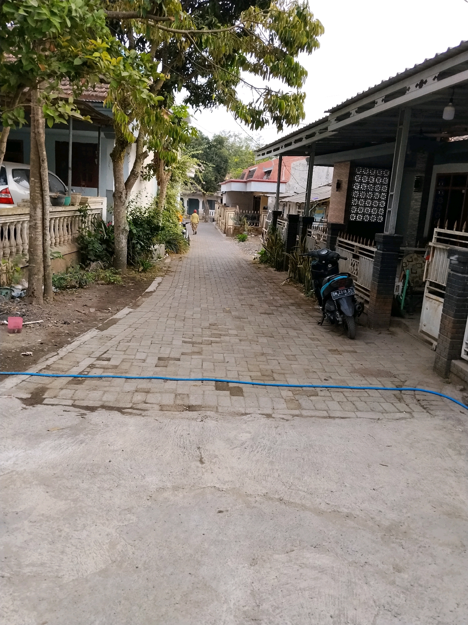 The image depicts a narrow residential street in Tlogosari, Indonesia. The street is paved with interlocking bricks, with a concrete sidewalk bordering one side. The scene is lined with houses, some with open carports and decorative fences. A white car is parked on the left side of the street. A person wearing a yellow shirt and a red headscarf is walking away from the camera down the street. A black motorcycle is parked on the right side of the street. The environment is lush with trees and vegetation, suggesting a tropical climate. A blue hose is laid across the street in the foreground. The sky is overcast, with a faint glow suggesting daylight.