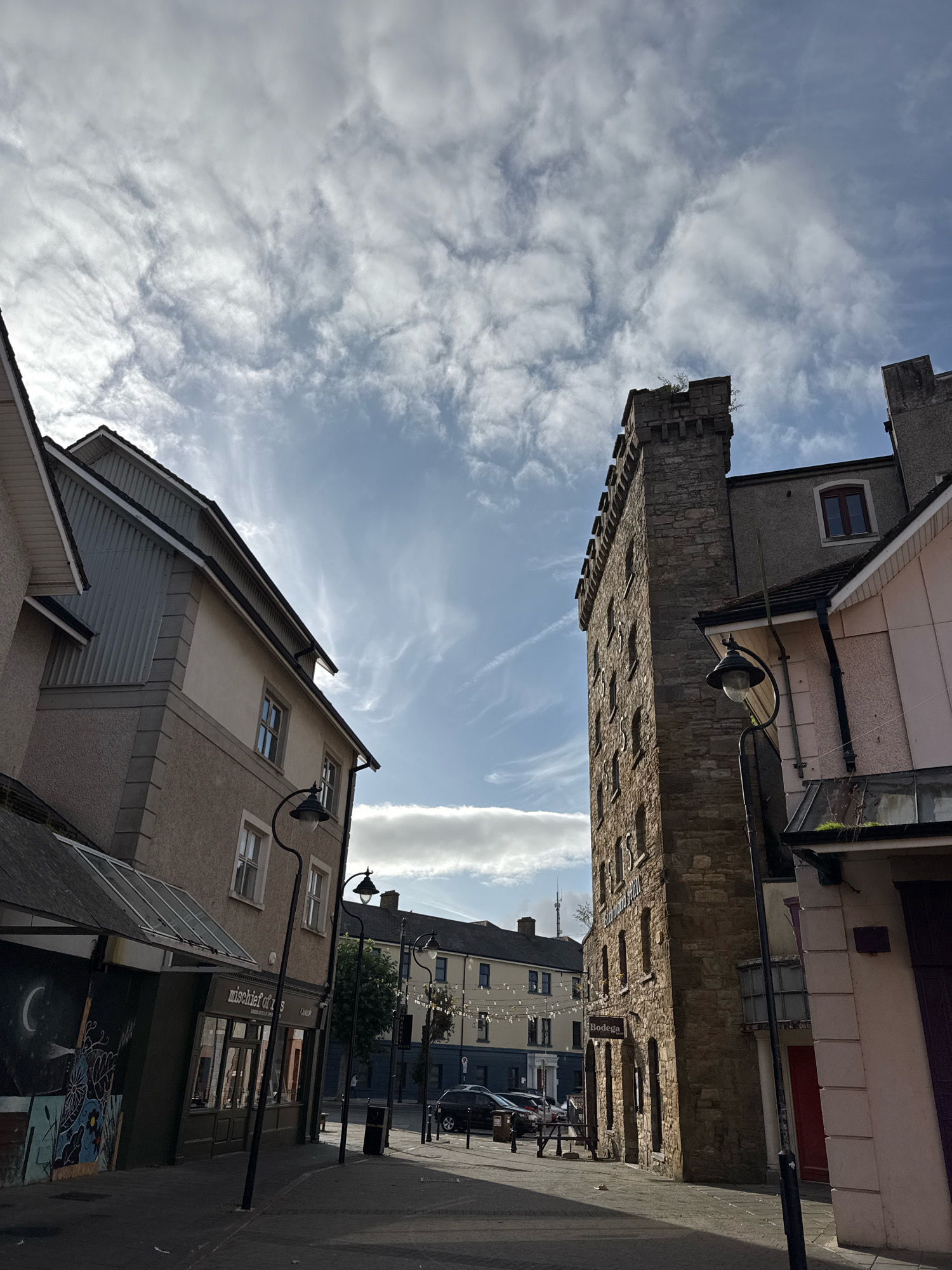 The image shows a street scene in Clonmel, Ireland. On the left is a building with a light-colored facade, dark grey ribbed siding on an upper section, and a black awning with a glass roof. Graffiti art featuring a crescent moon and floral designs is visible on the lower part of this building's wall. The building has several windows and a storefront with the text "mischief of 8" visible. Adjacent to this building on the right is a tall, narrow, stone building with a crenellated top, resembling a tower. This stone building has numerous small, arched windows. To the right of the stone building is a pink-colored structure with a dark doorway. Several street lamps with black poles and clear glass globes are positioned along the sidewalk. In the background, more buildings are visible, including ones with string lights overhead, and cars are parked on the street. The sky is a clear blue with scattered, wispy white clouds. The sun is casting shadows on the ground, indicating it is daytime. There are no people clearly visible in the foreground.