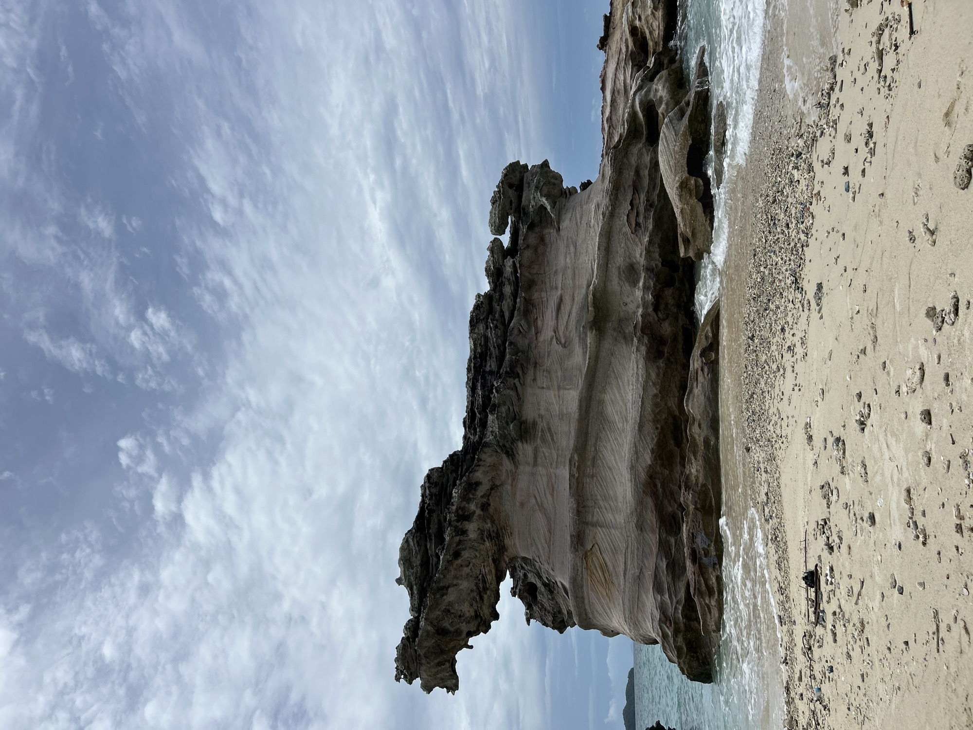 A large, sculpted rock formation is situated on the shore of a sandy beach in Phru Nai, Thailand. The rock, with its eroded and layered appearance, resembles a creature or a statue, with a prominent overhang on the left side that is reminiscent of a head. The rock is partially submerged in clear, turquoise water, with small waves lapping at its base. The beach is composed of fine sand and small pebbles, with scattered pieces of coral. The sky above is overcast with a pattern of layered clouds. The background shows a glimpse of another island in the distance. The scene is devoid of any people or discernible human activity.
