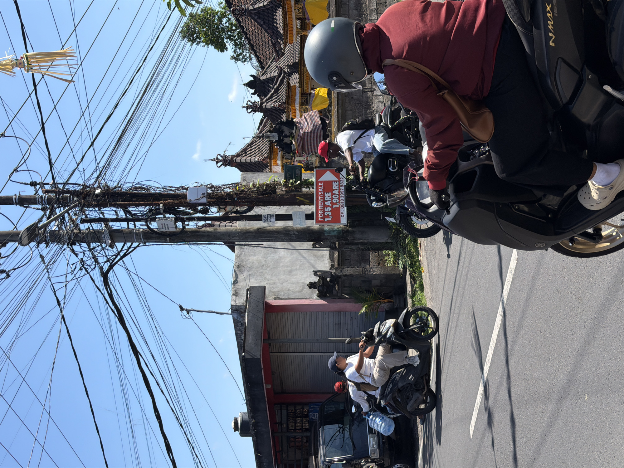 In the city of Mengwi, Indonesia, this image captures a street scene under a clear blue sky. A densely packed network of electrical wires crisscrosses the upper portion of the frame, suspended from a large utility pole. In the foreground, two individuals on motorcycles are traveling on a paved road. The person in the foreground, seen from the back, is wearing a maroon long-sleeved shirt, black pants, a dark grey helmet, and light-colored clogs. A brown shoulder bag is slung across their chest. The motorcycle they are riding is black and appears to be a scooter.
To the left, a second motorcycle is visible, carrying two people. The rider wears a white short-sleeved shirt, light-colored pants, a blue cap, and a backpack. The passenger, wearing a red baseball cap and a white shirt, is holding a large clear plastic bottle of water. Their motorcycle is also black.
Behind the motorcycles, a building with a grey concrete facade and a pink trim is situated. It has large, rolled-down metal shutters. To the right of this building, traditional Balinese-style roof structures are visible, adorned with yellow and red fabric banners, suggesting a cultural or religious setting. A sign with red text on a white background, reading "FOR RENT/SALE 1,35 ARE 1.90 ARE CP: 0813 3997 8822", is attached to a pole. The overall environment suggests a busy urban street with a blend of modern infrastructure and local architectural elements.