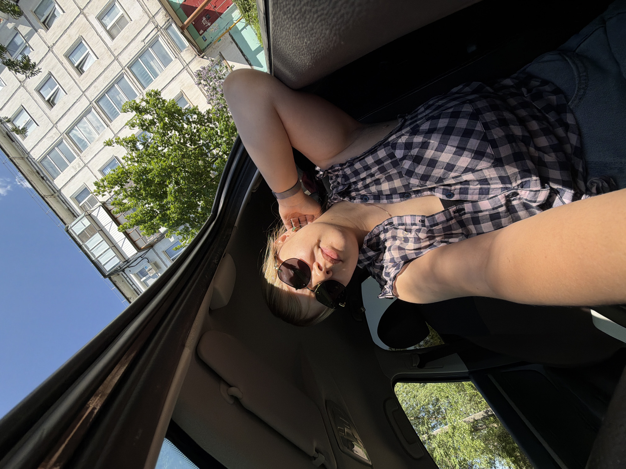 A young woman with fair skin and blonde hair is inside a car. She is wearing dark sunglasses and a plaid shirt. The car is parked in a location that appears to be Chaikovsky, Russia, judging by the architecture visible through the car window. In the background, there is a light-colored apartment building with multiple windows, along with trees and a blue sky. The photo is taken from a low angle within the car, looking up and slightly sideways at the woman.