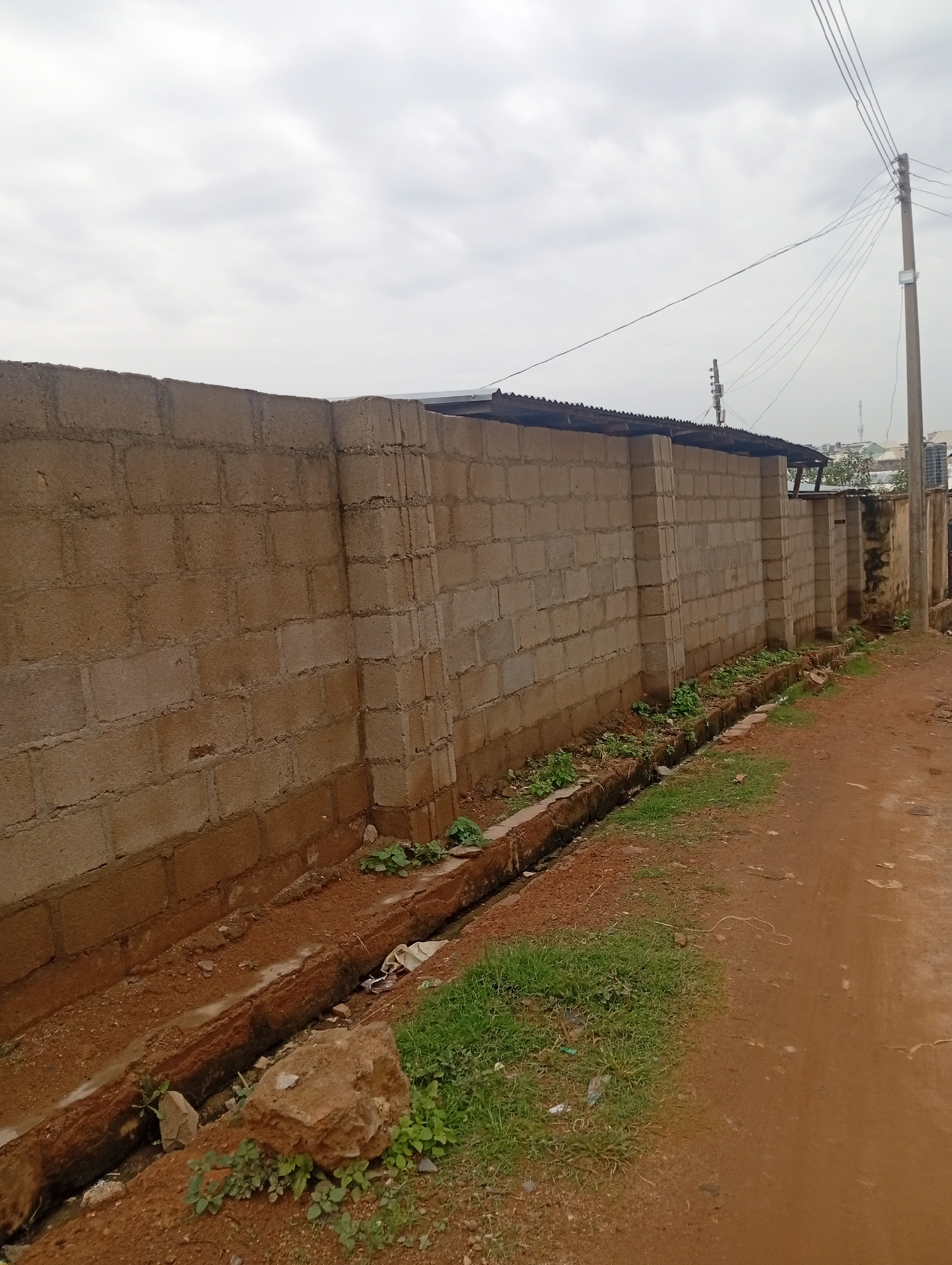 The image depicts a scene in the city of Rafin Par, Nigeria. A weathered, light brown cinder block wall stretches across the frame, forming a boundary. The wall is constructed with horizontal blocks and vertical supports, showing signs of age and exposure to the elements. To the left of the wall, a narrow, shallow drainage ditch runs along its base, filled with water and debris. Some green plants are growing in and around the ditch, with a large rock visible in the foreground. The ground beside the ditch is a reddish-brown dirt road, with patches of dry grass. Overhead, a cloudy sky prevails, and a utility pole with several wires is visible on the right side of the frame, extending into the sky. In the distance, some buildings and structures can be seen.