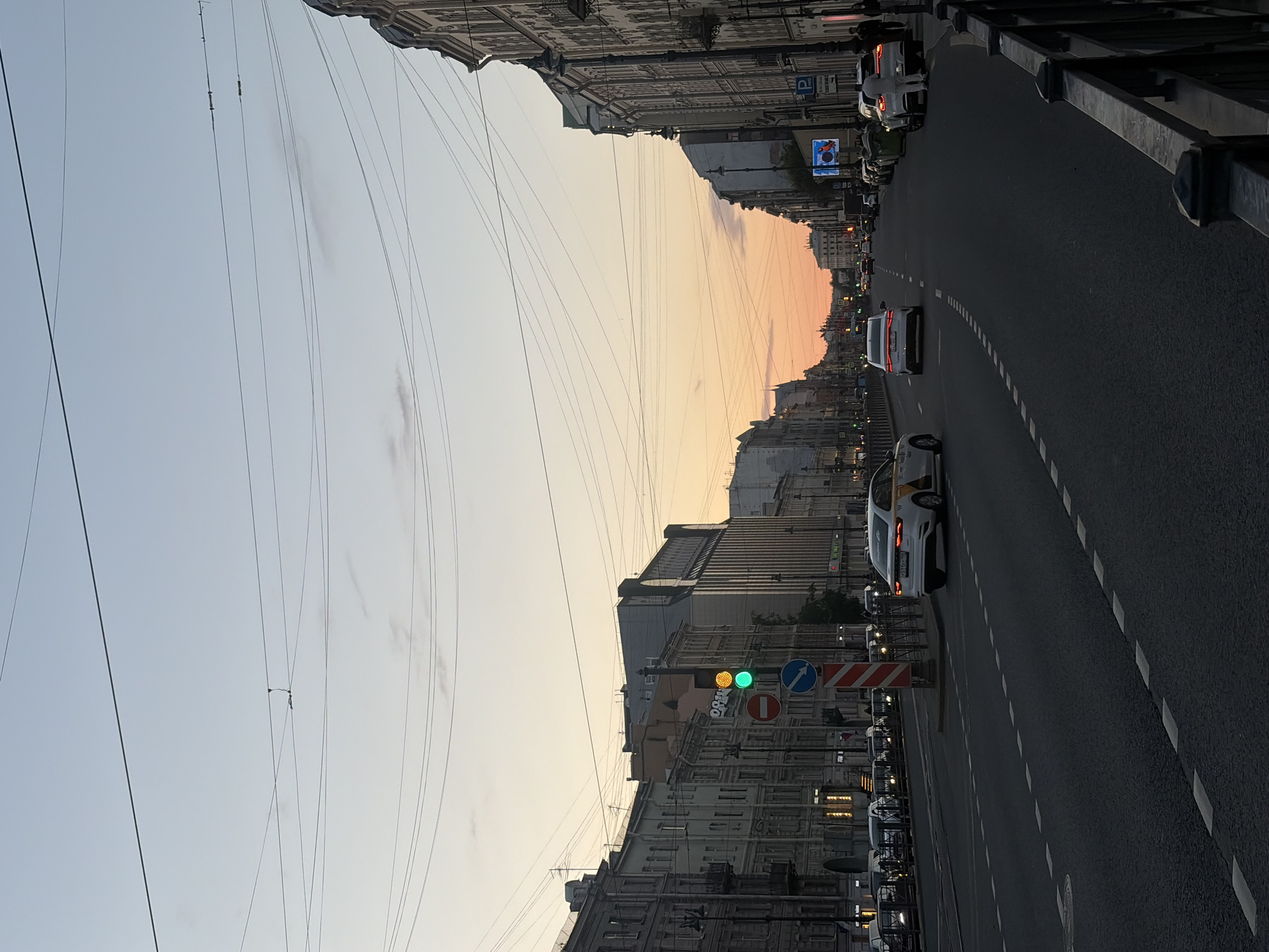 This is a medium shot of a street in Saint Petersburg, Russia, during twilight. The sky is a gradient of pale blue and soft orange. A wide asphalt road with white dashed lane markers dominates the foreground. Several cars are driving on the road, including a white taxi with yellow and black markings on the side. On the left side of the road, a traffic light displays green and yellow. Buildings with ornate architecture line both sides of the street. Overhead, a complex network of electrical wires crisscrosses the sky. Pedestrians are visible on the sidewalks, and a black metal fence runs along the right side of the road. A parking sign is visible on the right side of the street.