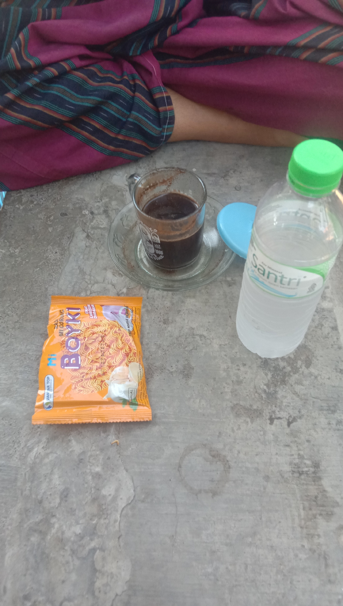 In a setting possibly in or near Ngempit, Indonesia, a concrete surface displays various objects. A person's legs, covered by a sarong with dark blue, green, and maroon stripes, are visible in the upper left portion of the image. In the center, a glass cup filled with dark liquid, likely coffee, rests on a saucer. To the right of the coffee cup is a clear plastic bottle of water with a green cap and a label that reads "Santri." In the foreground, an orange packet of instant noodles, labeled "Boyki Mi Goreng," is laid flat. The overall scene suggests a casual moment of refreshment.
