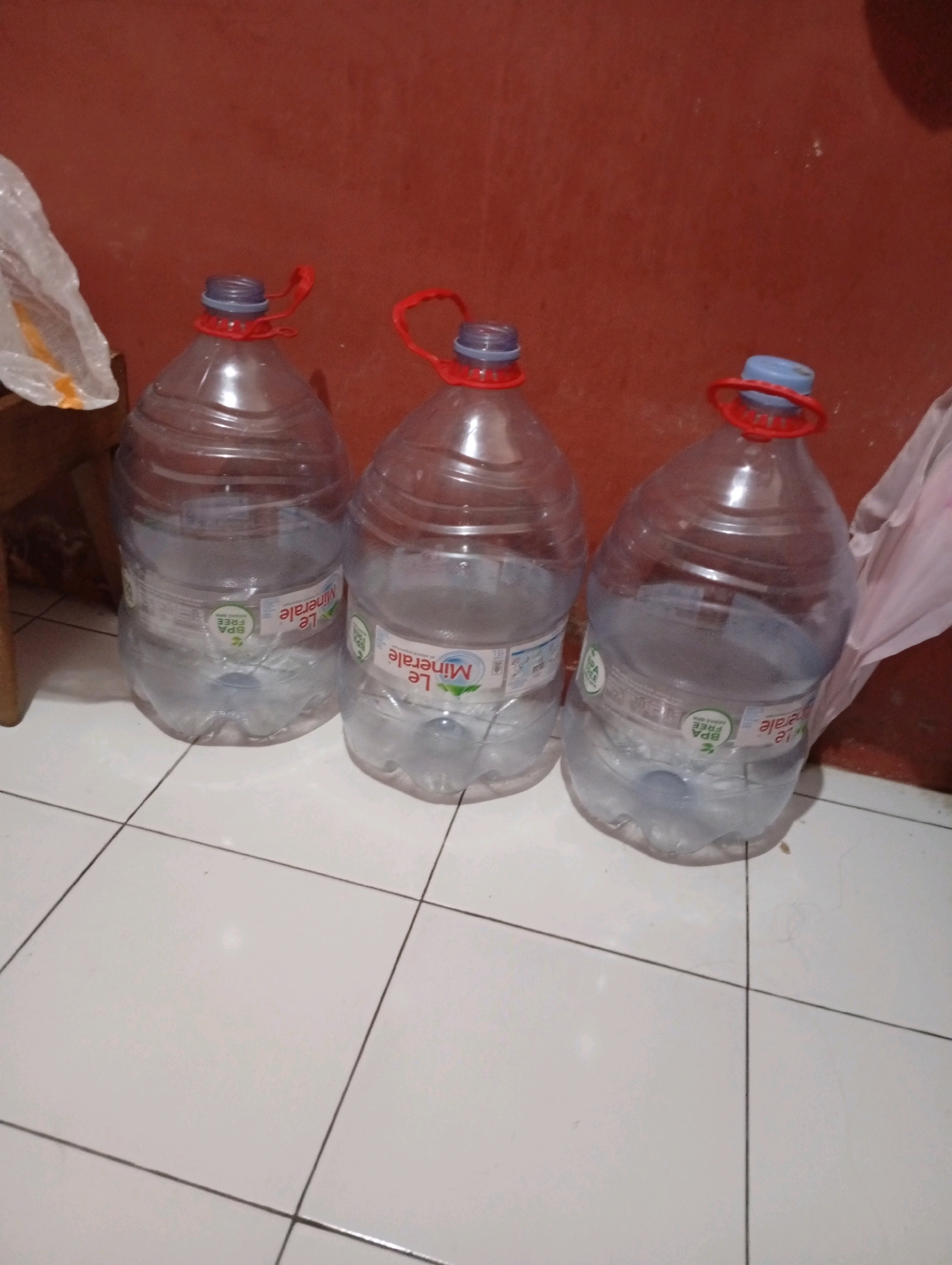 Three large, clear plastic water bottles with blue caps and red carrying handles are arranged in a row on a tiled floor. The bottles appear to be partially filled with water. Each bottle has a label that reads "Le Minerale" and some other text that is not fully legible, including "BPA FREE". The background is a reddish-brown wall. A white plastic bag is visible to the left of the bottles. The scene is indoors, likely in a residential setting in Tasikmalaya, Indonesia.