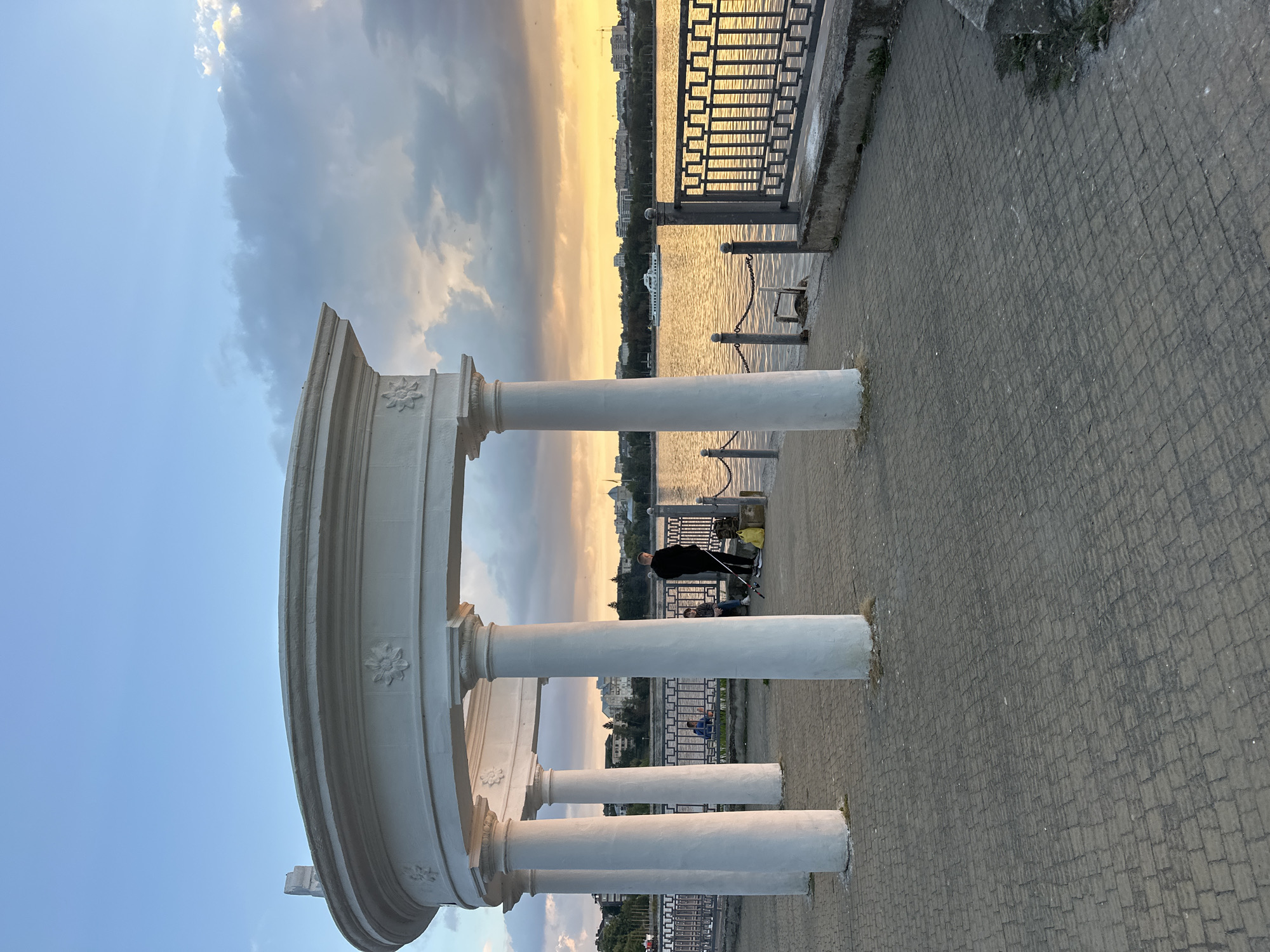 The image shows a sunset scene at the Ternopil lake promenade in Ternopil, Ukraine. A white, curved, open-air structure with classical columns stands on a paved walkway. In the foreground, a person in dark clothing stands near a yellow bag and fishing rod, seemingly fishing. To the left, another person is seated near a railing. The lake, reflecting the warm colors of the sunset, is visible in the background, with a white boat sailing on its surface. The cityscape of Ternopil is visible on the opposite bank of the lake, with buildings and trees lining the shore. The sky is a mix of blue and dramatic clouds tinged with orange and yellow from the setting sun.