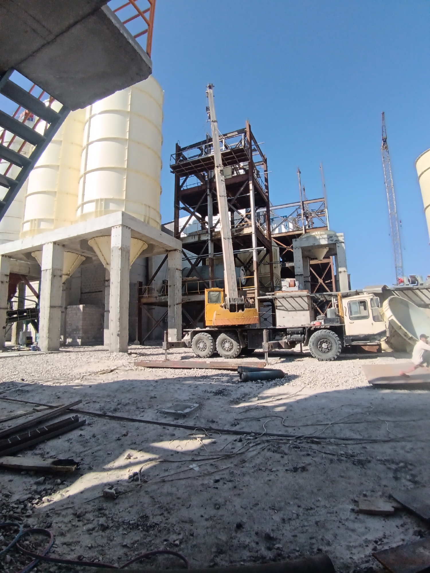 The image depicts an outdoor construction site with industrial equipment under a clear blue sky. The scene is dominated by large, cream-colored silos supported by concrete pillars on the left side. In the center and right, a multi-story industrial structure is under construction, made of metal beams and girders. A yellow crane truck is positioned in front of this structure, with its boom extended upwards. Adjacent to the truck is another large piece of industrial equipment, possibly a concrete mixer or silo component, with a wide opening. The ground is covered with gravel, construction debris, and metal pipes. A figure, partially visible on the far right, appears to be interacting with the equipment. The overall impression is one of ongoing construction or industrial activity.