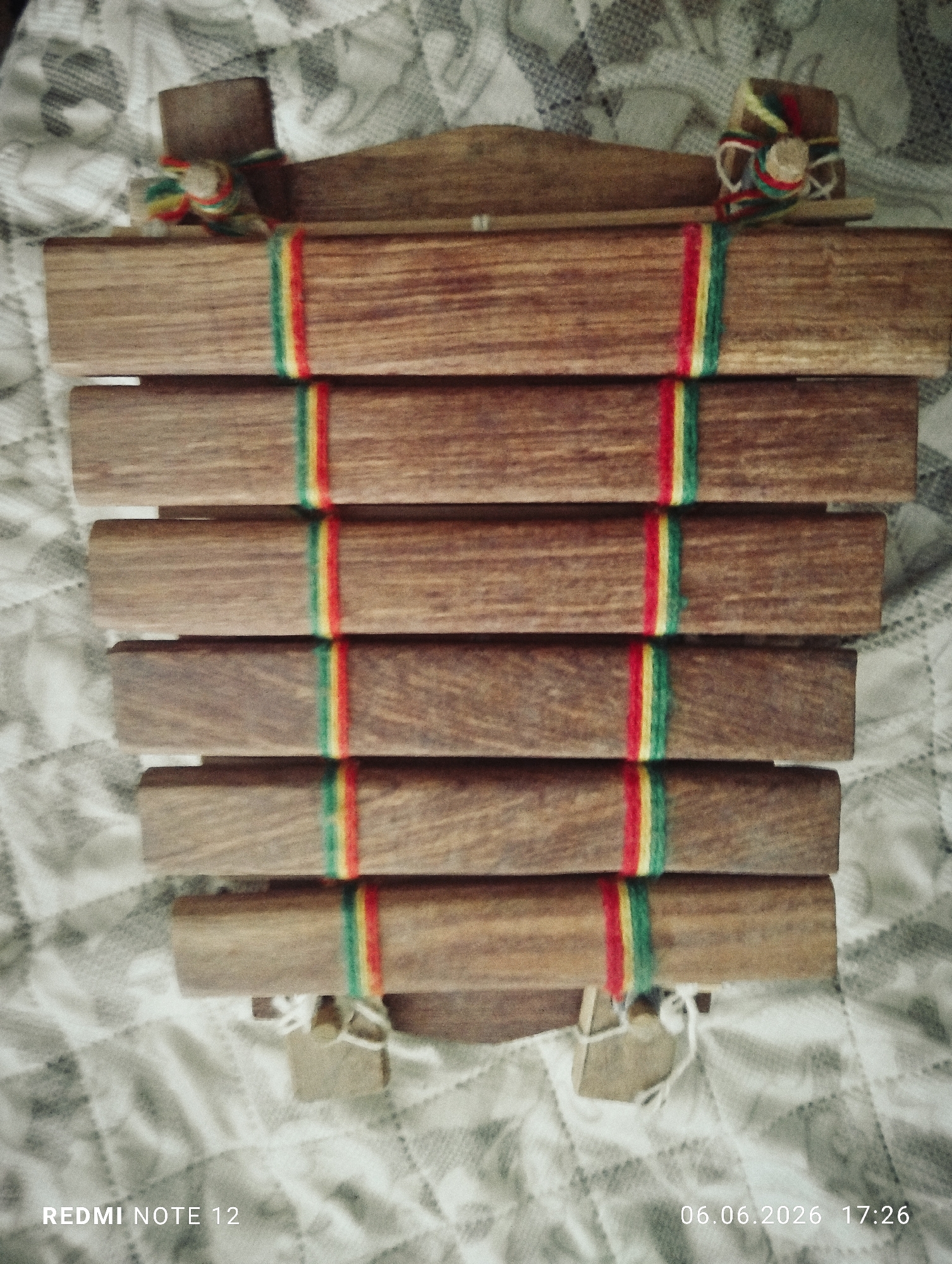 The image displays a portable wooden musical instrument, resembling a xylophone or balafon, resting on a patterned textile surface. The instrument features six horizontal wooden bars of varying lengths, arranged in a descending sequence from top to bottom. Each bar is a medium-brown tone with visible wood grain. A vertical strip of multi-colored yarn or string, composed of red, yellow, and green bands, is wrapped around the center of each wooden bar, creating a consistent visual element across the instrument.
The wooden bars are supported by a frame structure. At the upper end, two small vertical wooden blocks are present, each adorned with a small, bundled decoration of red, yellow, and green string. A thin, light-colored wooden stick, consistent with a mallet, rests horizontally across the top section of the instrument. The lower supports of the instrument also show white string or cord material.
The background consists of a light-colored fabric with a repetitive, muted grey and white geometric or quilted pattern. The image exhibits soft, even illumination. Digital text overlays include "REDMI NOTE 12" in the lower-left corner and the timestamp "06.06.2026 17:26" in the lower-right corner. The image does not contain any discernible visual cues indicating the location in the city of Burgas, Bulgaria.