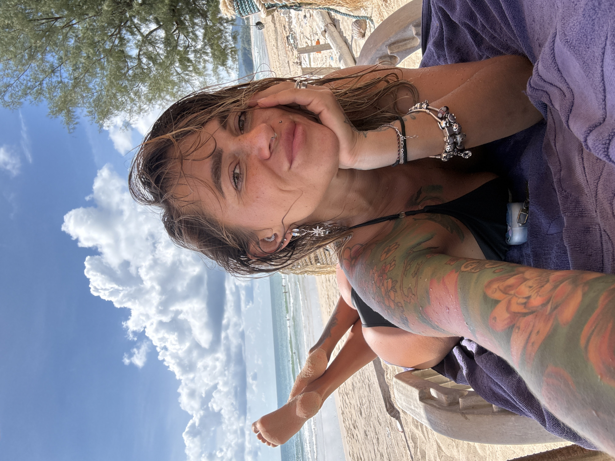 A woman with wet hair and tattoos on her arm is lying on a lounge chair on a sandy beach. She is wearing a black bikini top and has her hand resting on her chin. She has a nose ring and earrings. The ocean and a cloudy blue sky are visible in the background. The scene appears to be set during the daytime with warm weather, as indicated by the beach setting and the woman's attire. The overall mood seems relaxed and peaceful. The image does not contain any visible text.